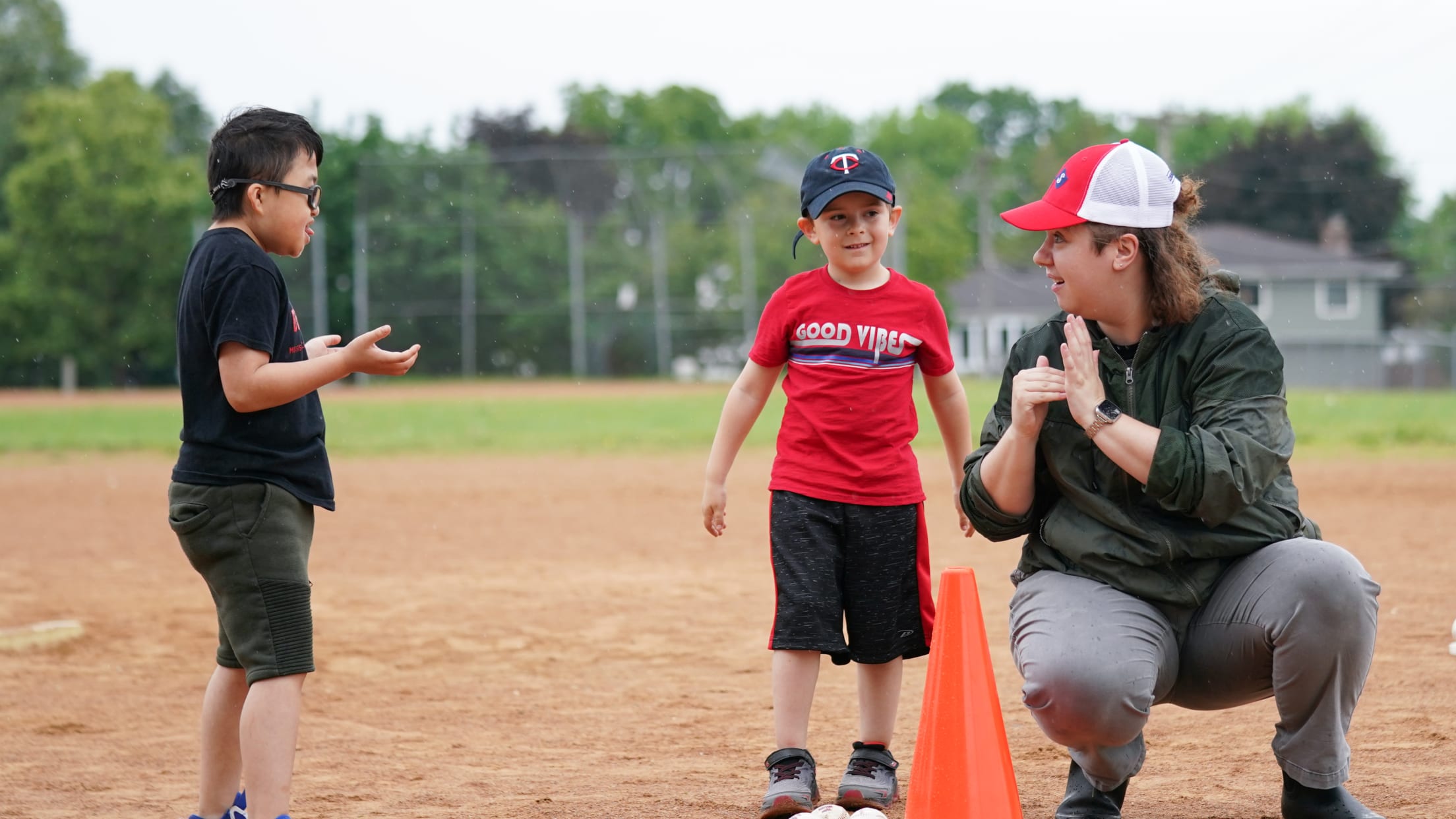 Two boys stand on a ballfield next to an adult volunteer, who is squatting down. The adult volunteer is communicating with the boys using American Sign Language.