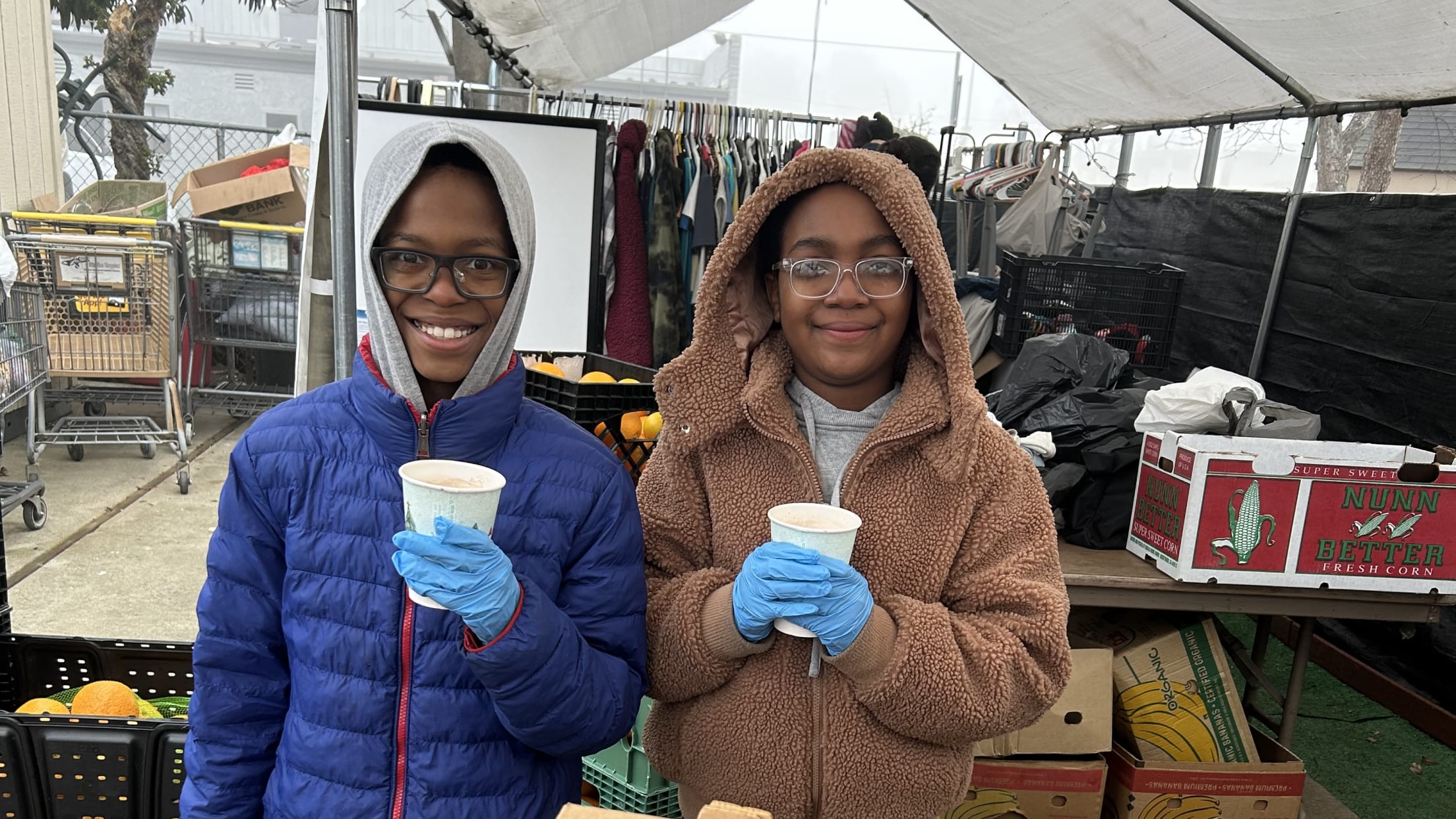 Two volunteers stand outside in front of a box of tomatoes with hot beverages in their hands.