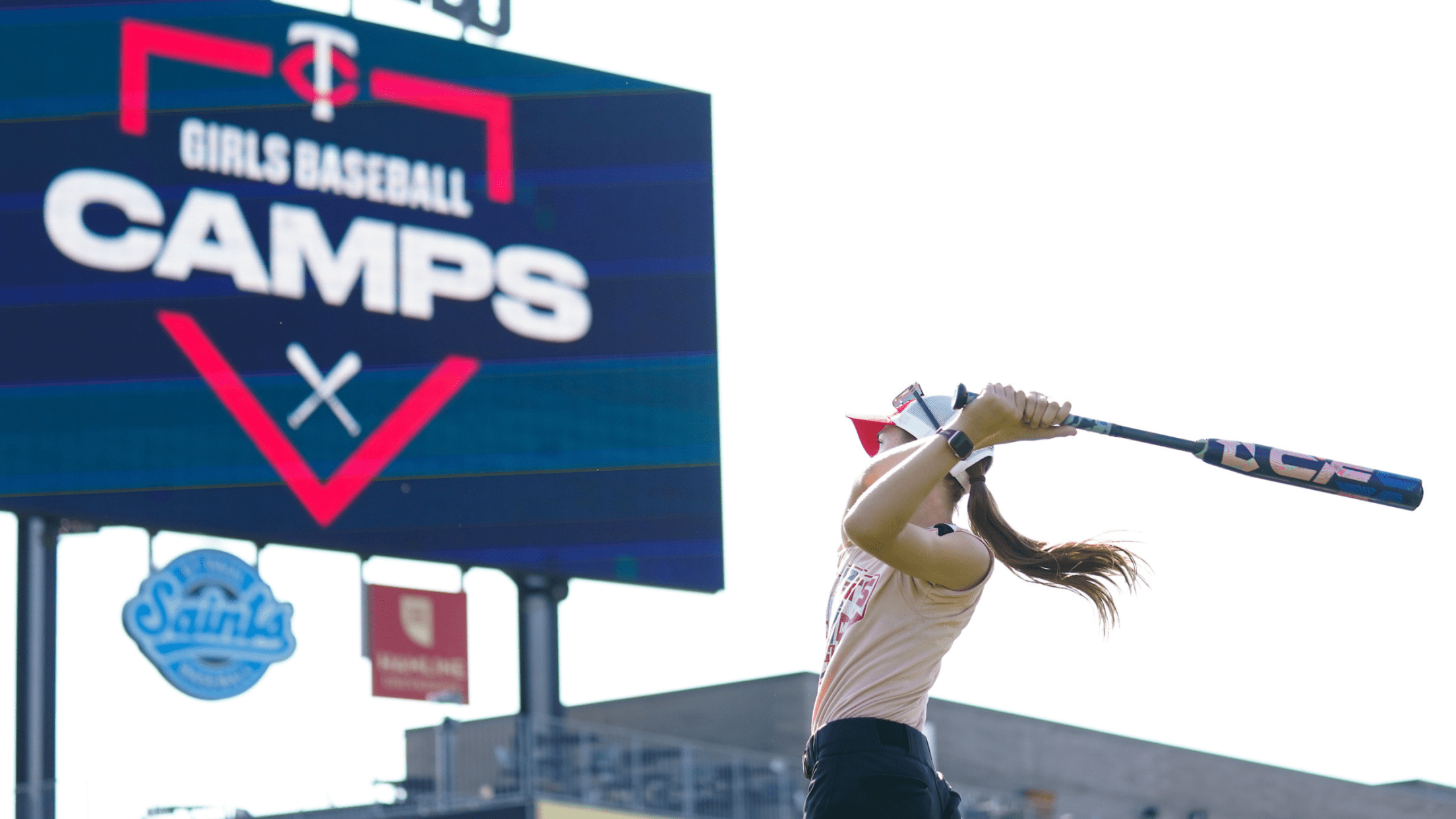Girl swinging bat. In the background is a videoboard with the Twins Girls Baseball Camps logo displayed.