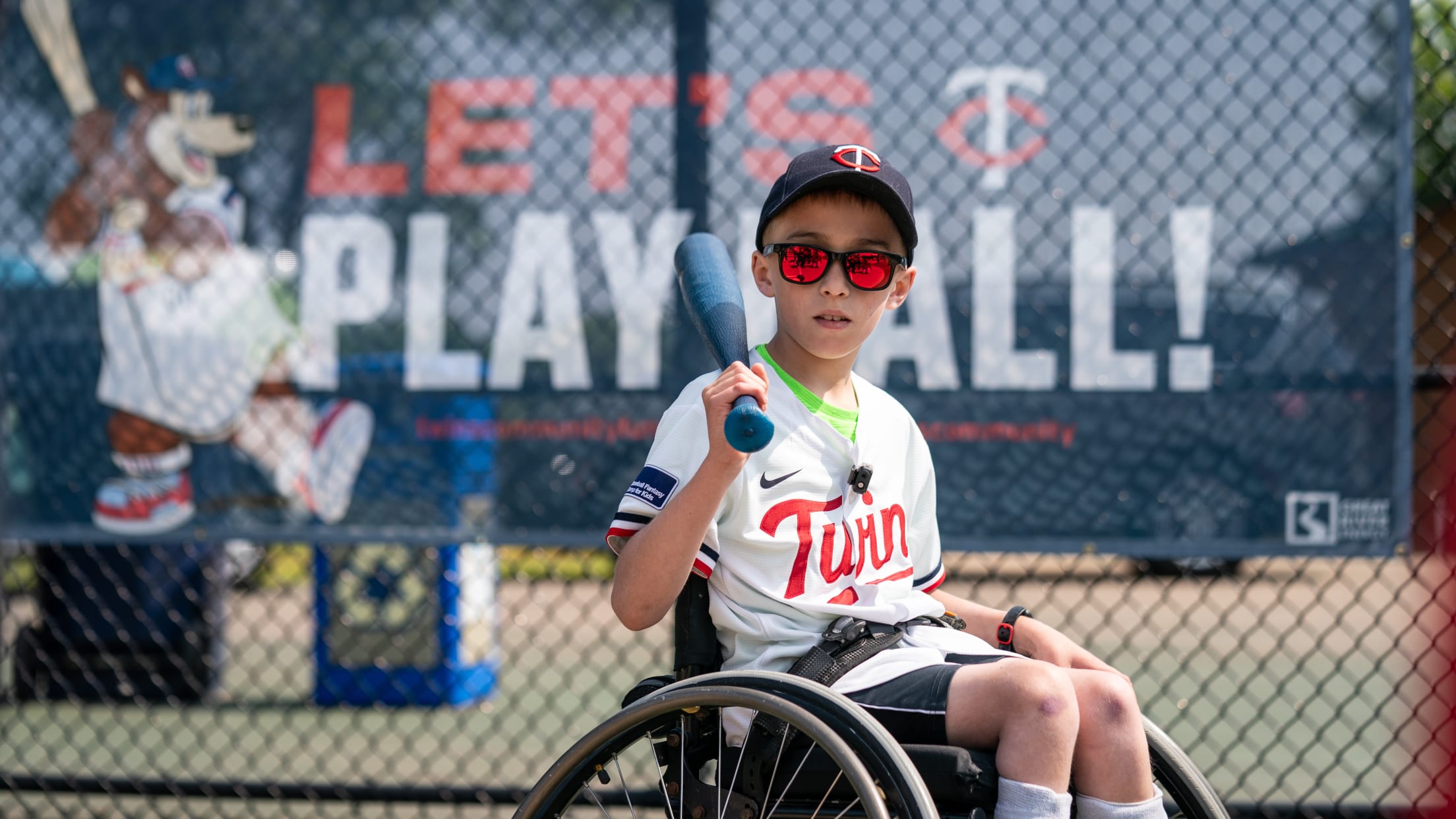 A boy using a wheelchair and wearing a Minnesota Twins jersey holds a bat on his shoulder while positioned in front of a banner that says "Let's Play Ball!"