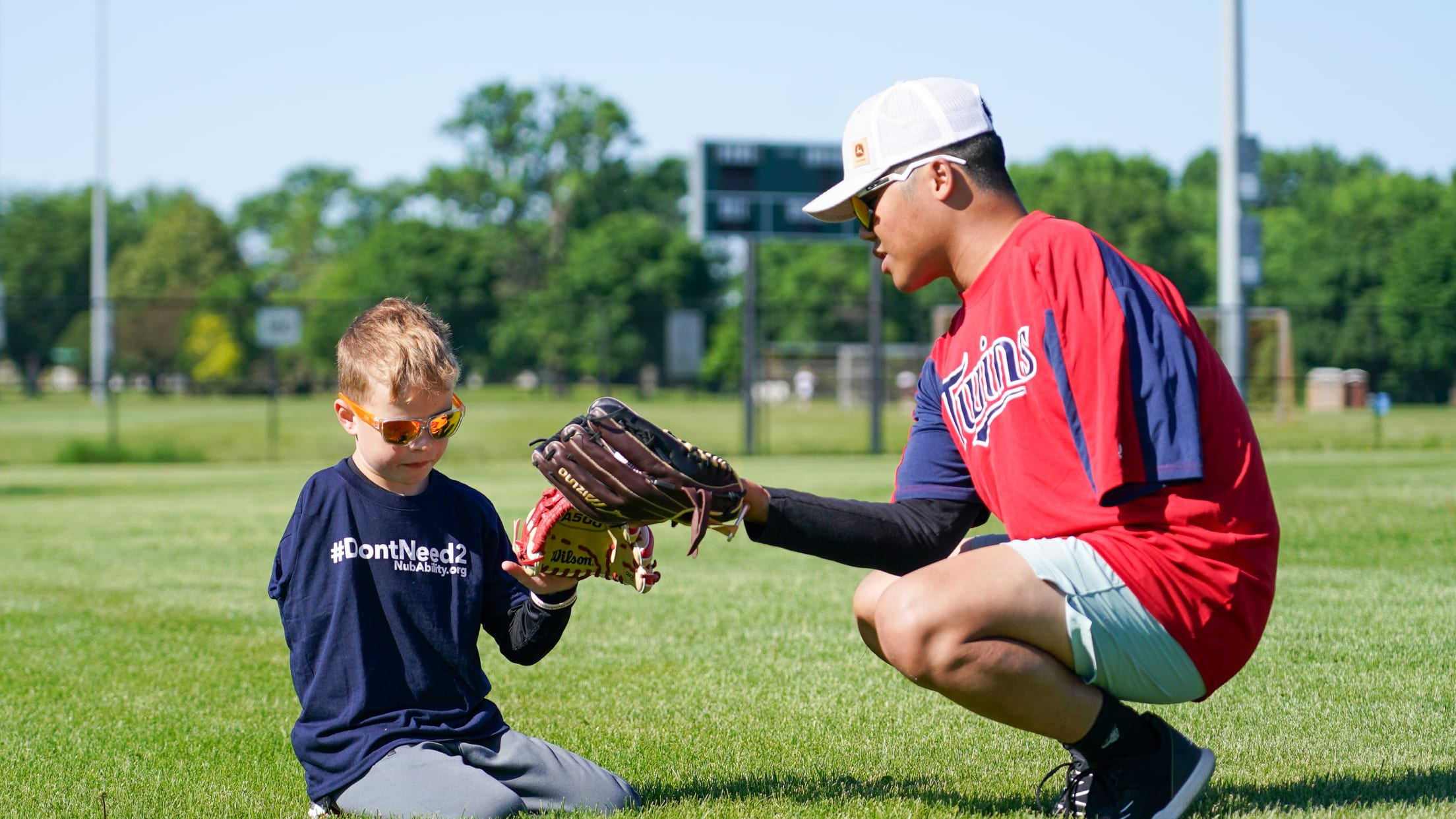 A limb-different boy kneels in the grass holding a baseball glove. A limb-different adult coach squats next to the boy showing him how to hold his glove.