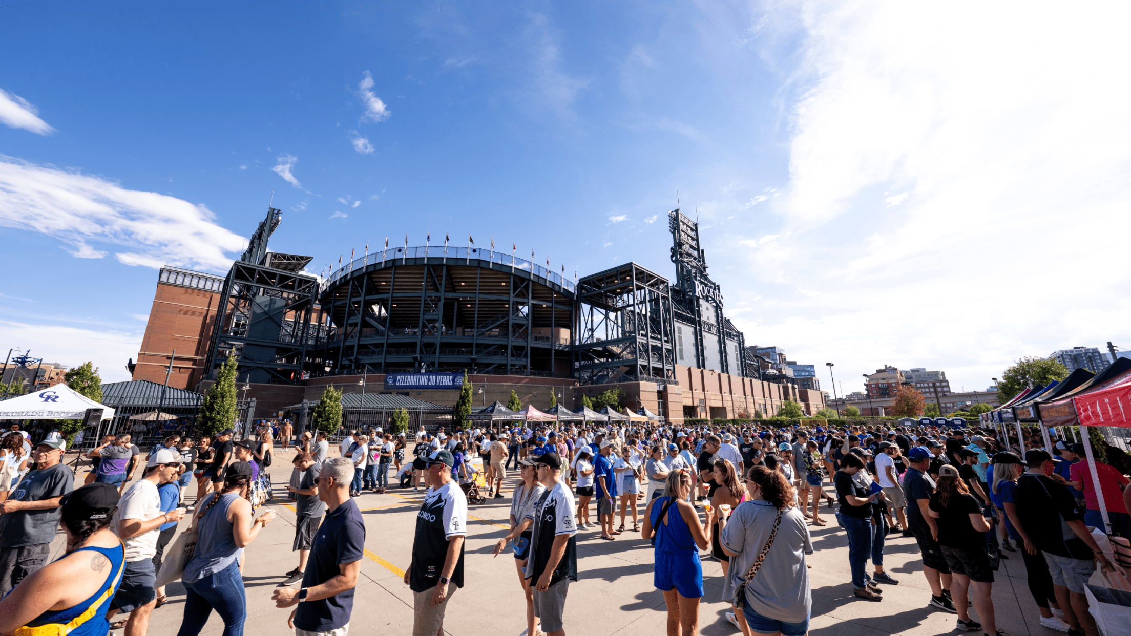 Parking lot with pop up tents on the outside, full of people standing in line and talking to each other with view of Coors Field ballpark in the background