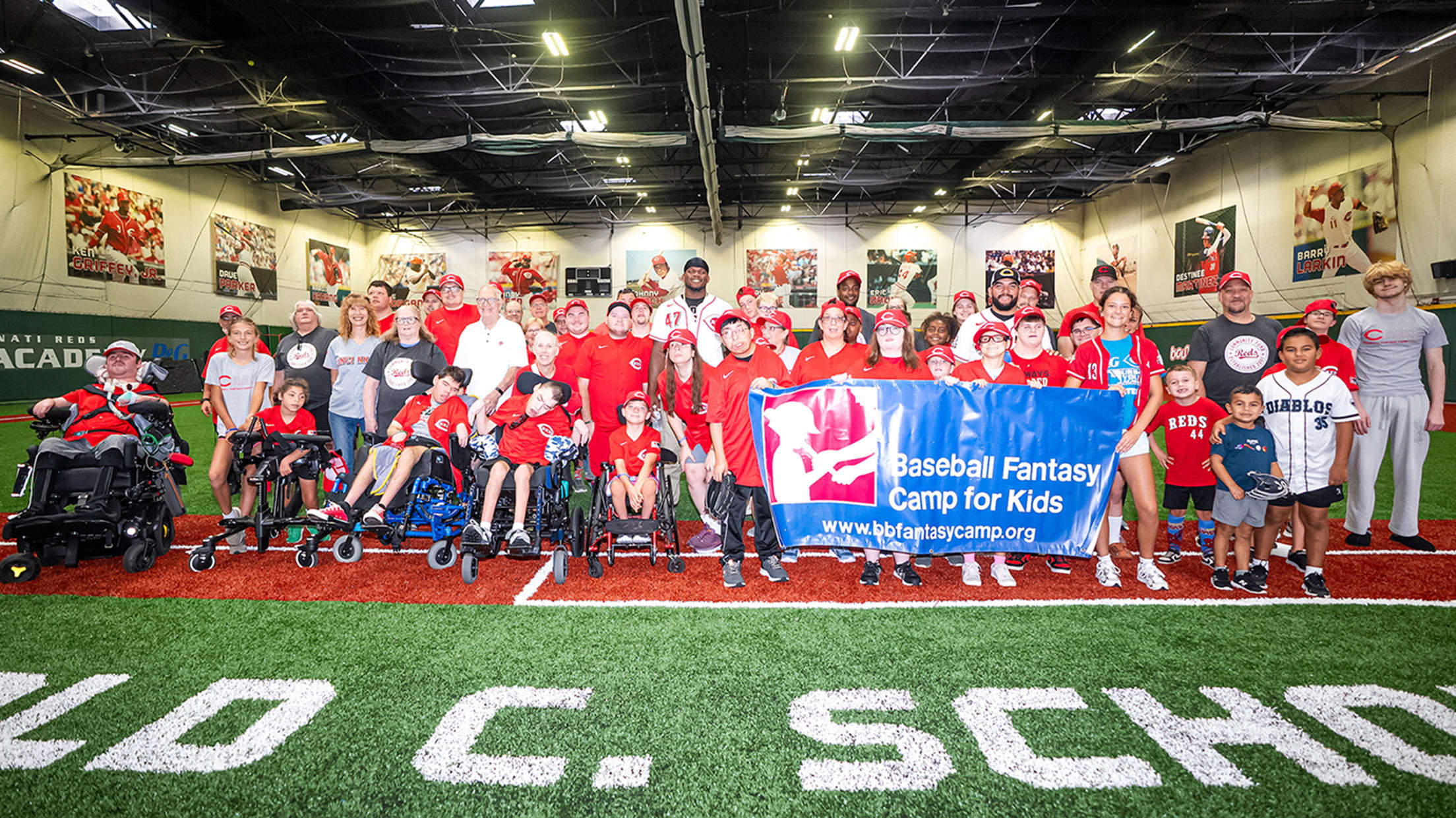 The kids and Reds players posing with the "Baseball Fantasy Camp for Kids" banner at the Reds Youth Academy.