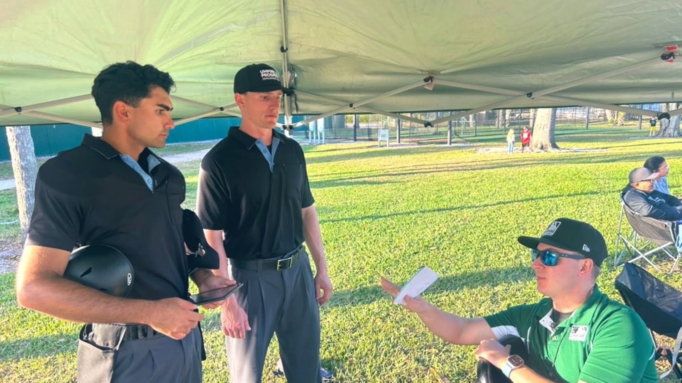 Two umpire prospects in uniform stand as a man in a green polo speaks to them.