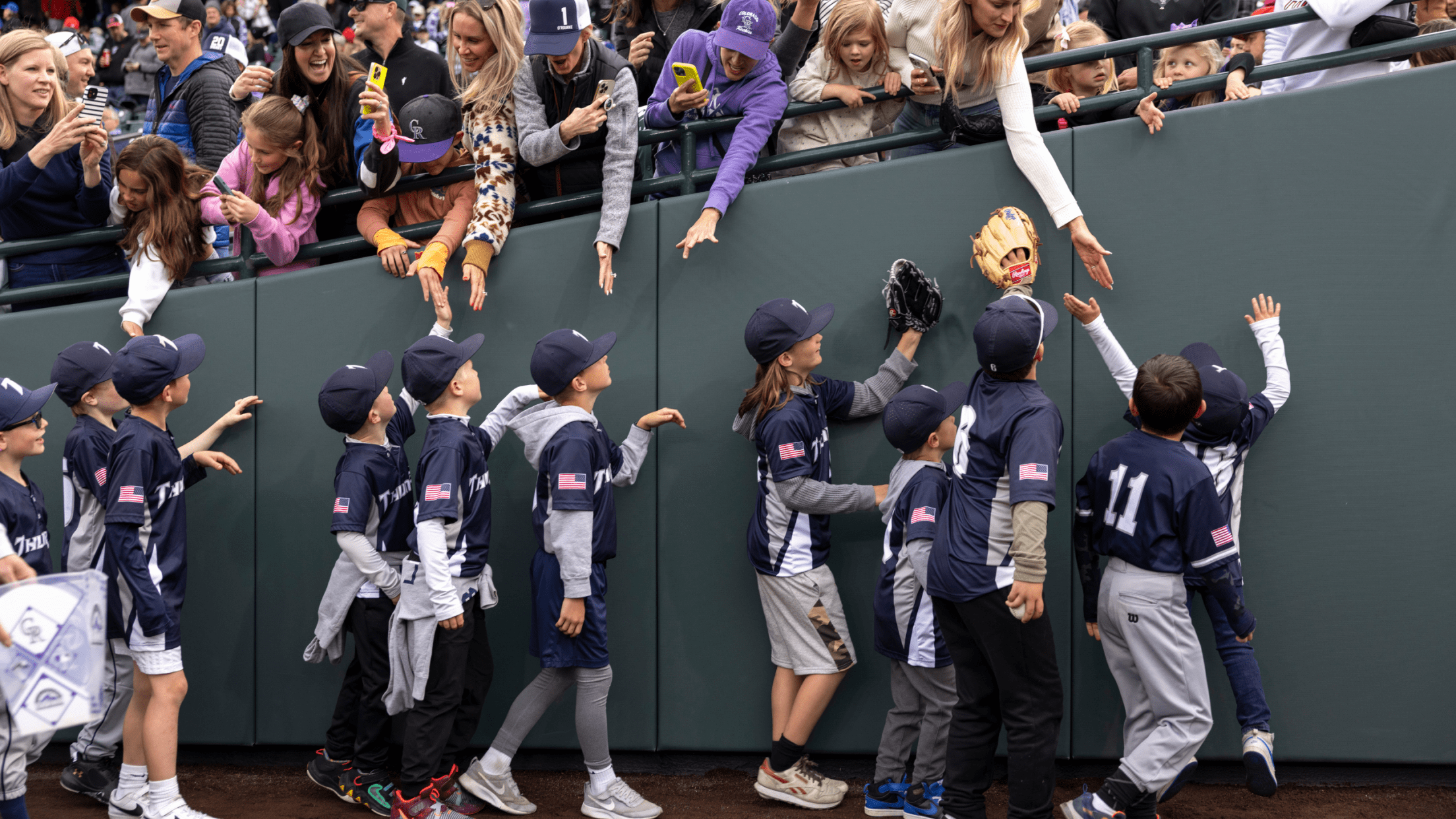 Youth baseball players walking on the Warning Track at Coors Field with fans in the stands taking pictures