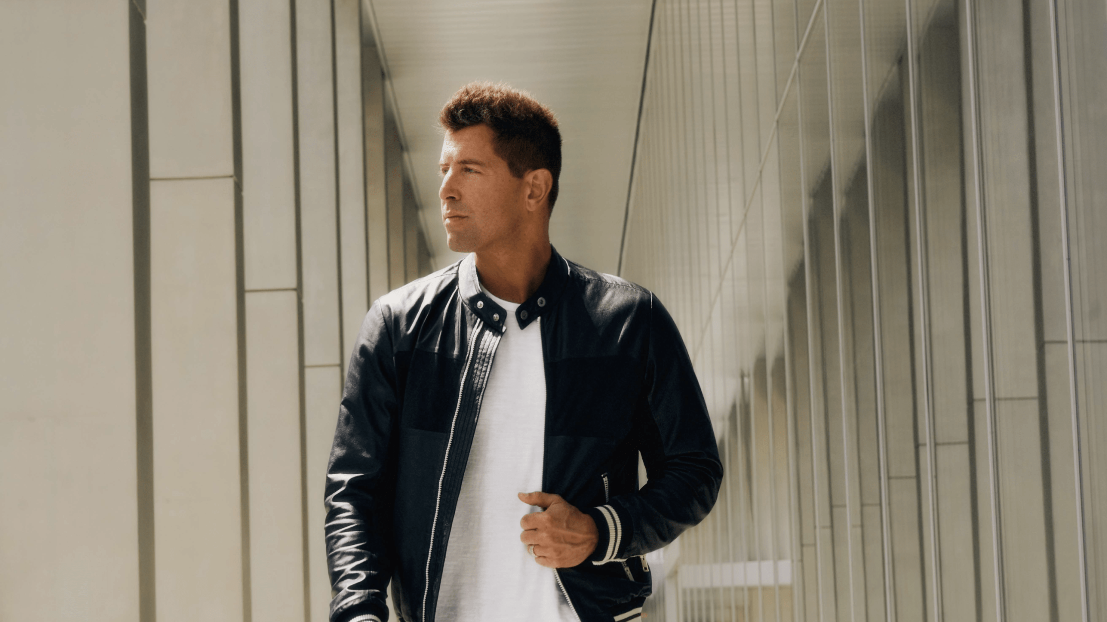 Male in white shirt and black jacket walking towards camera and looking left with concrete columns in background