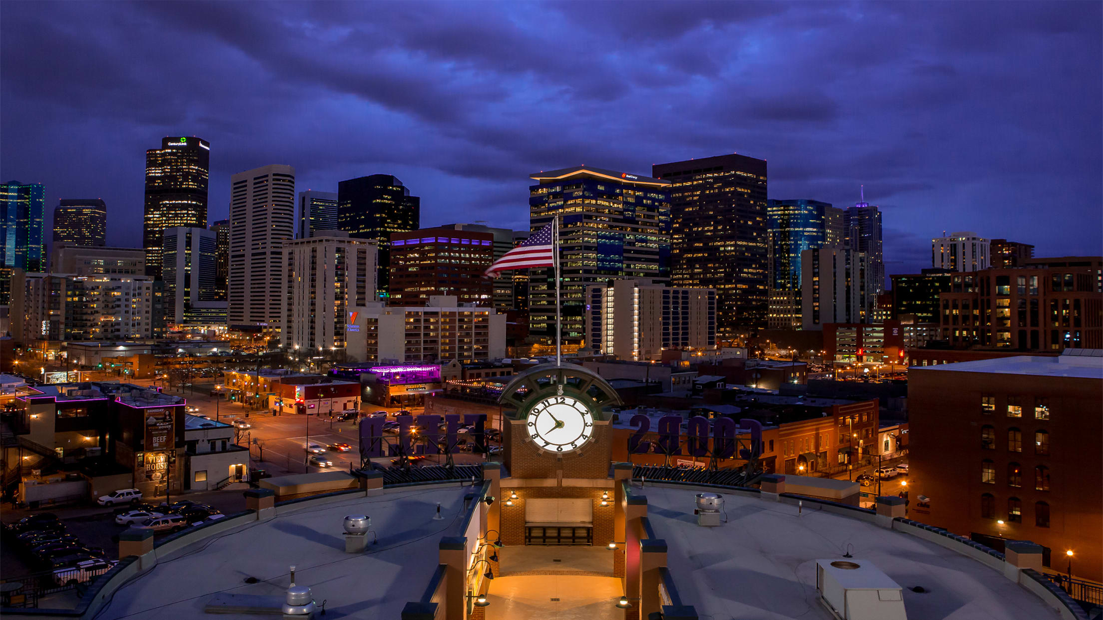 Downtown Denver skyline at dusk with illuminated city buildings, iconic clock tower, and American flag waving above LoDo district rooftops