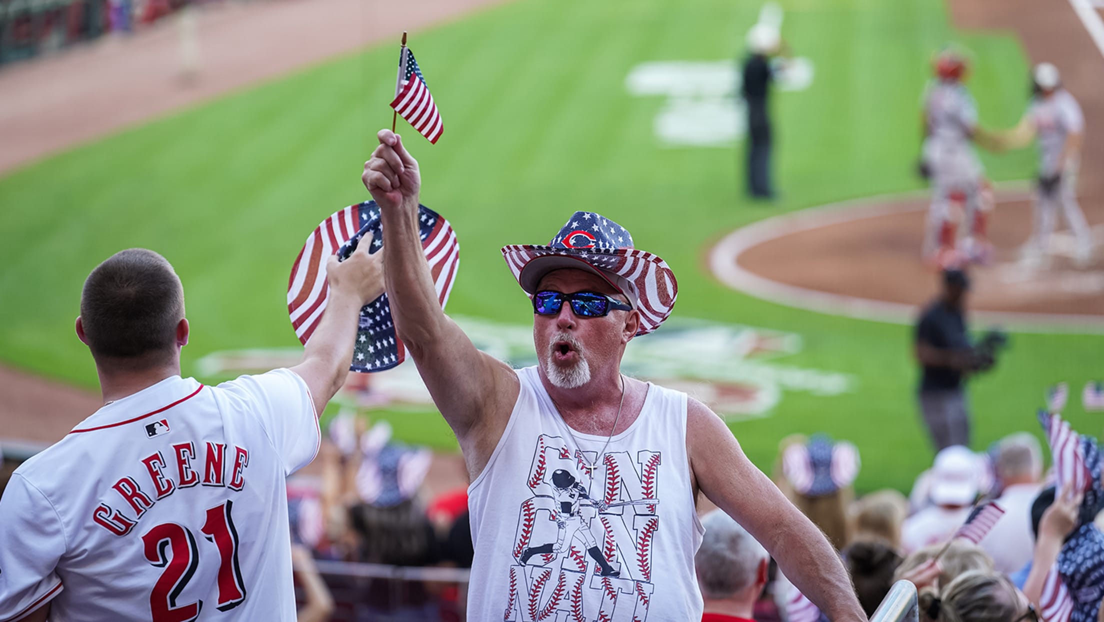 Reds fan wearing USA-themed cowboy hat and waving mini American flag during a special in-game moment.