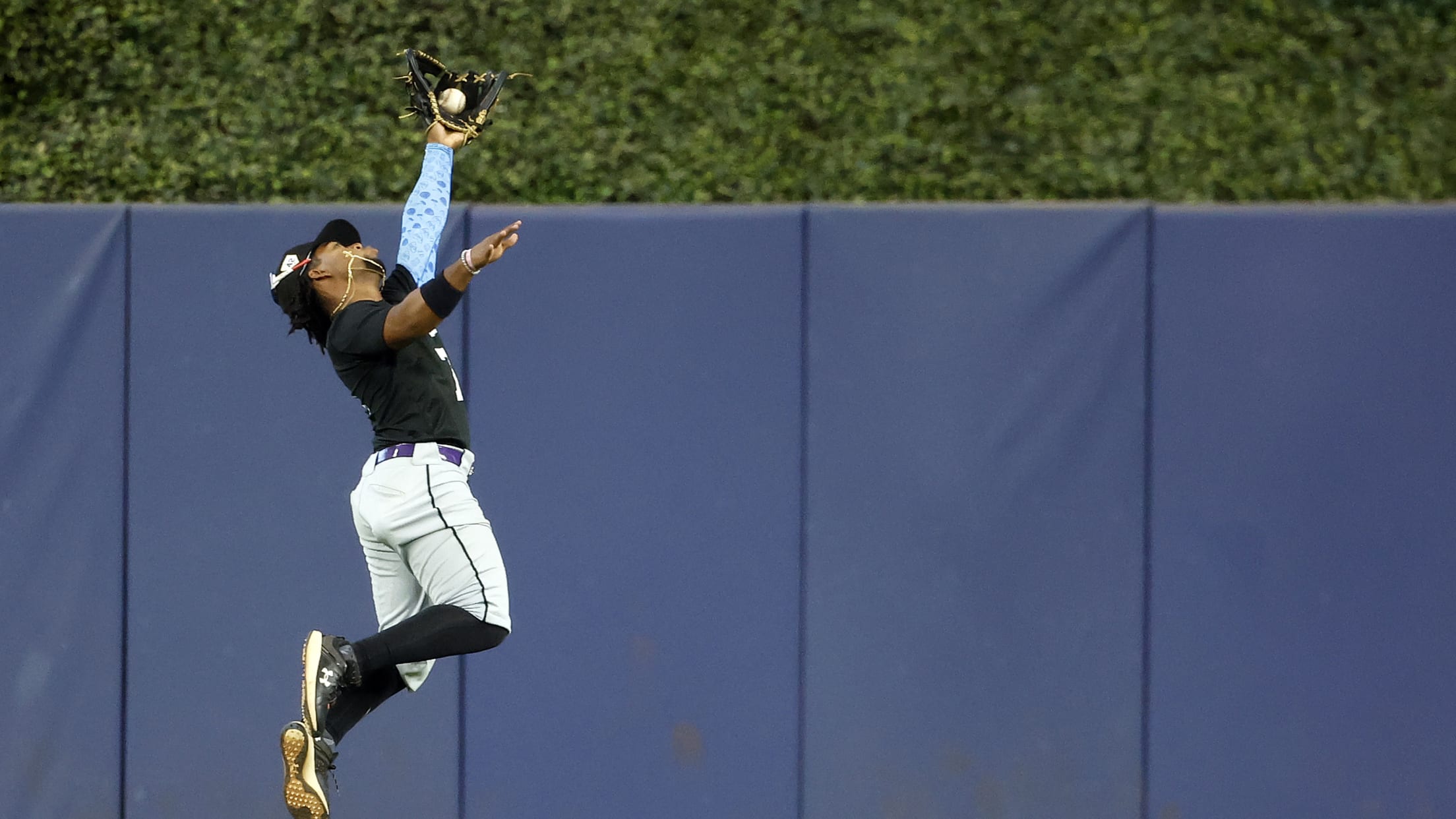 The outfielder jumps high in the air to catch the ball in front of the outfield wall.