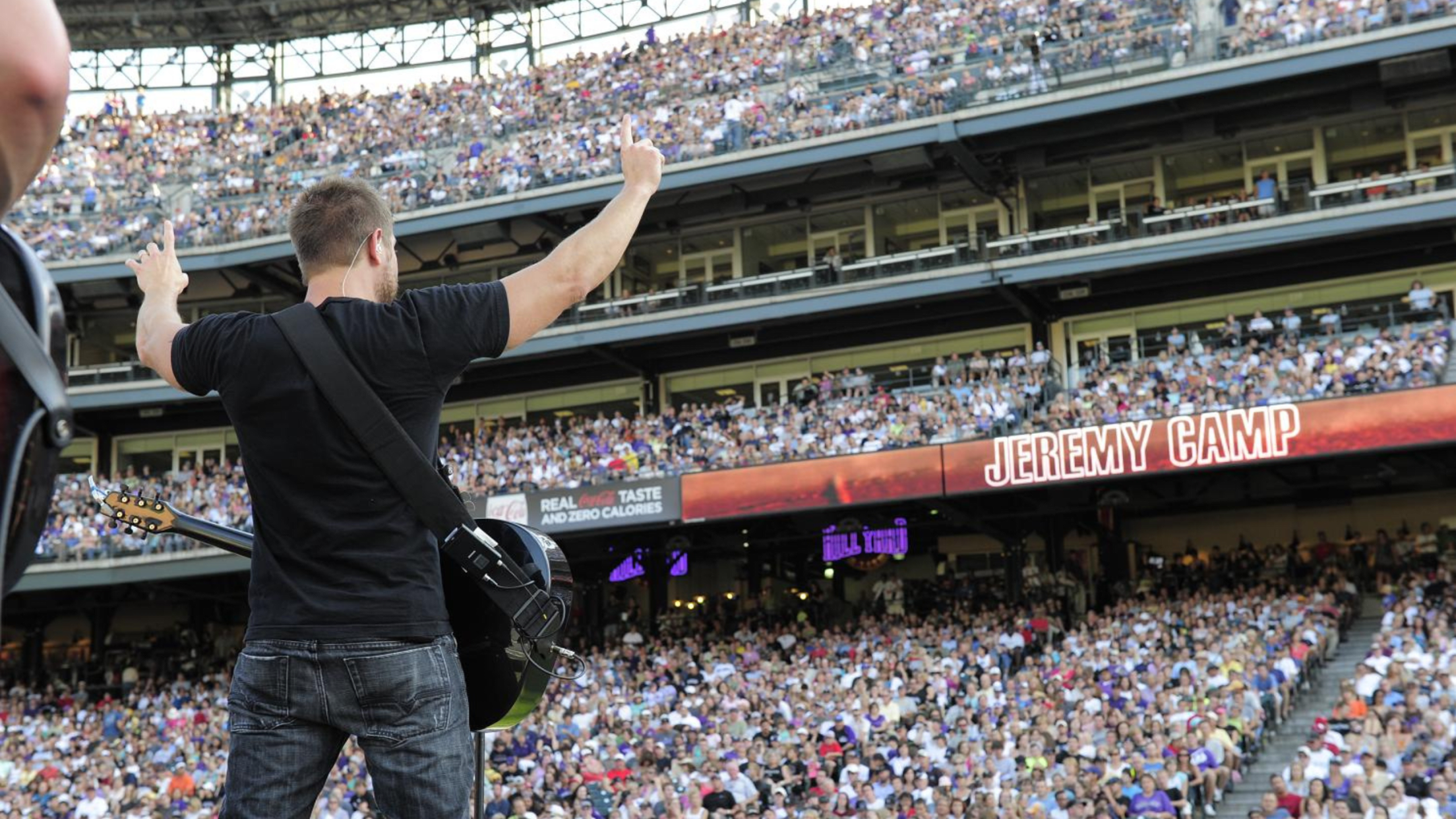 Male performer on stage with guitar facing crowd in stands at Coors Field