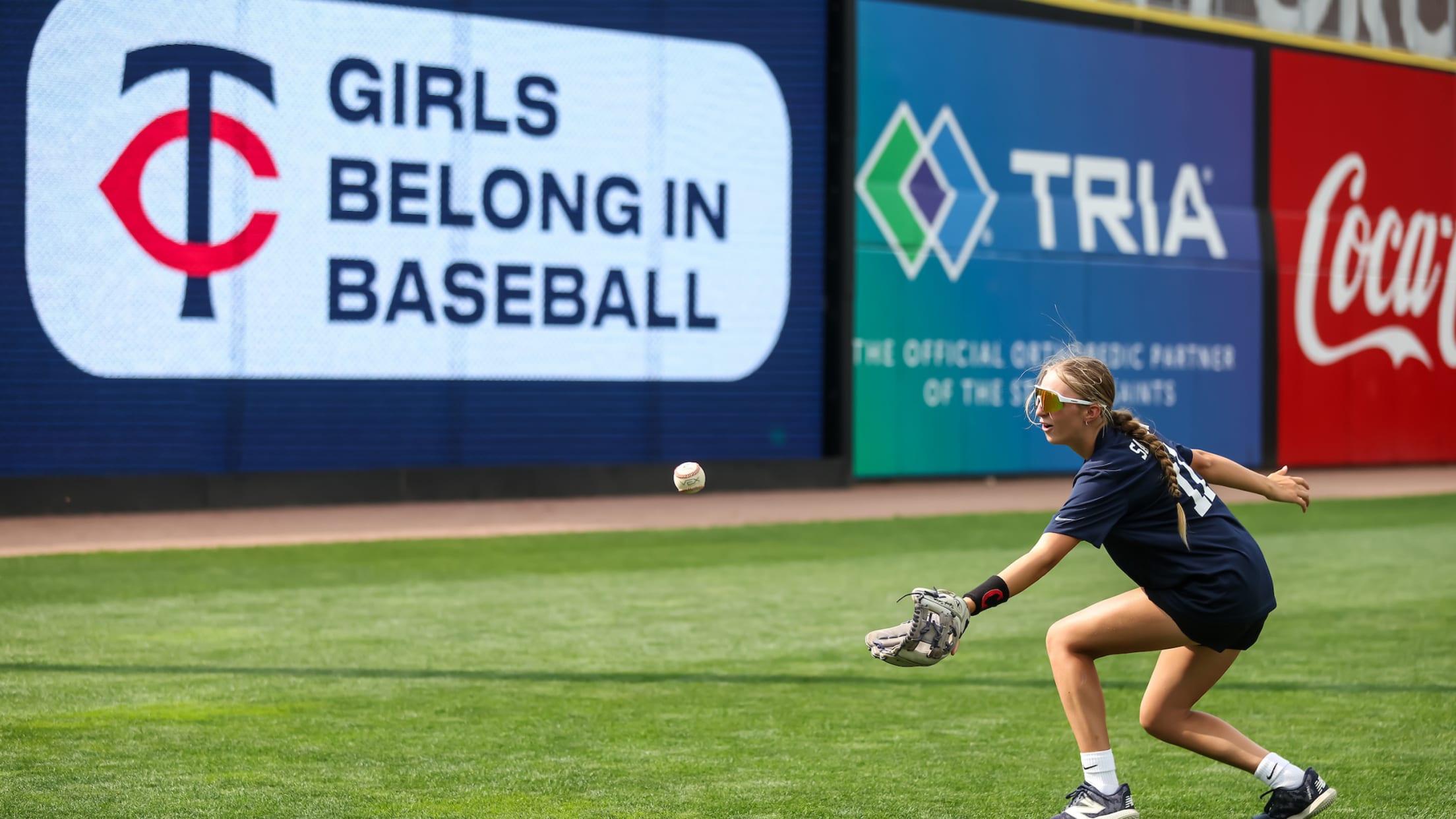 Girl catching baseball at Target Field. In the background a sign includes the Twins logo and text next to it that says 'girls belong in baseball'