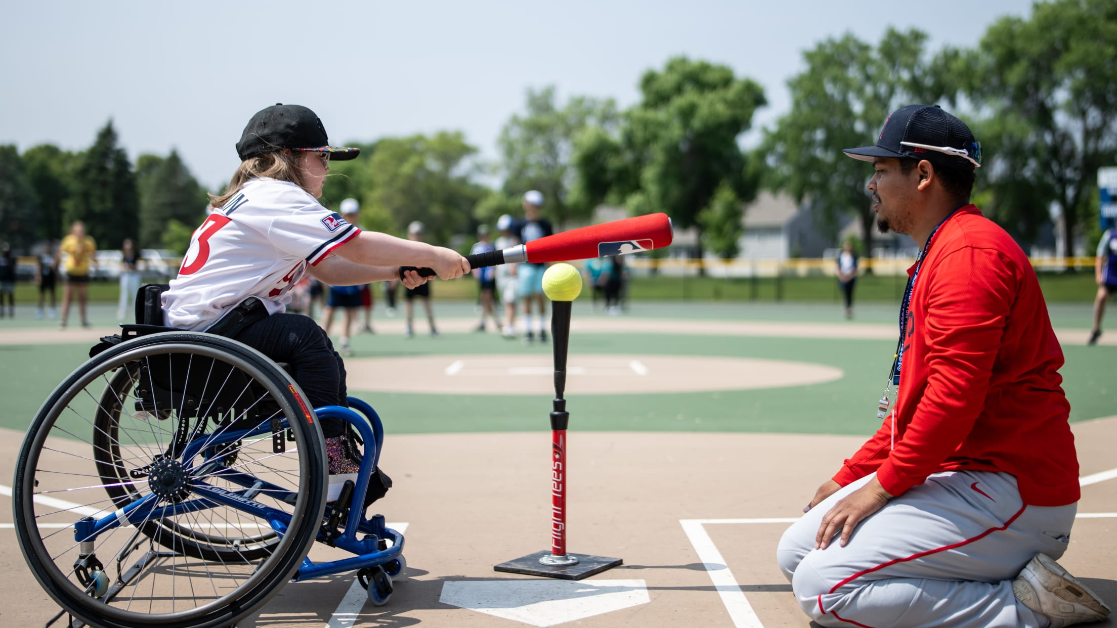 A girl using a wheelchair and wearing a Minnesota Twins jersey holds a bat and prepares to hit a ball off of a tee positioned in front of her. An adult coach is kneeling across from her.