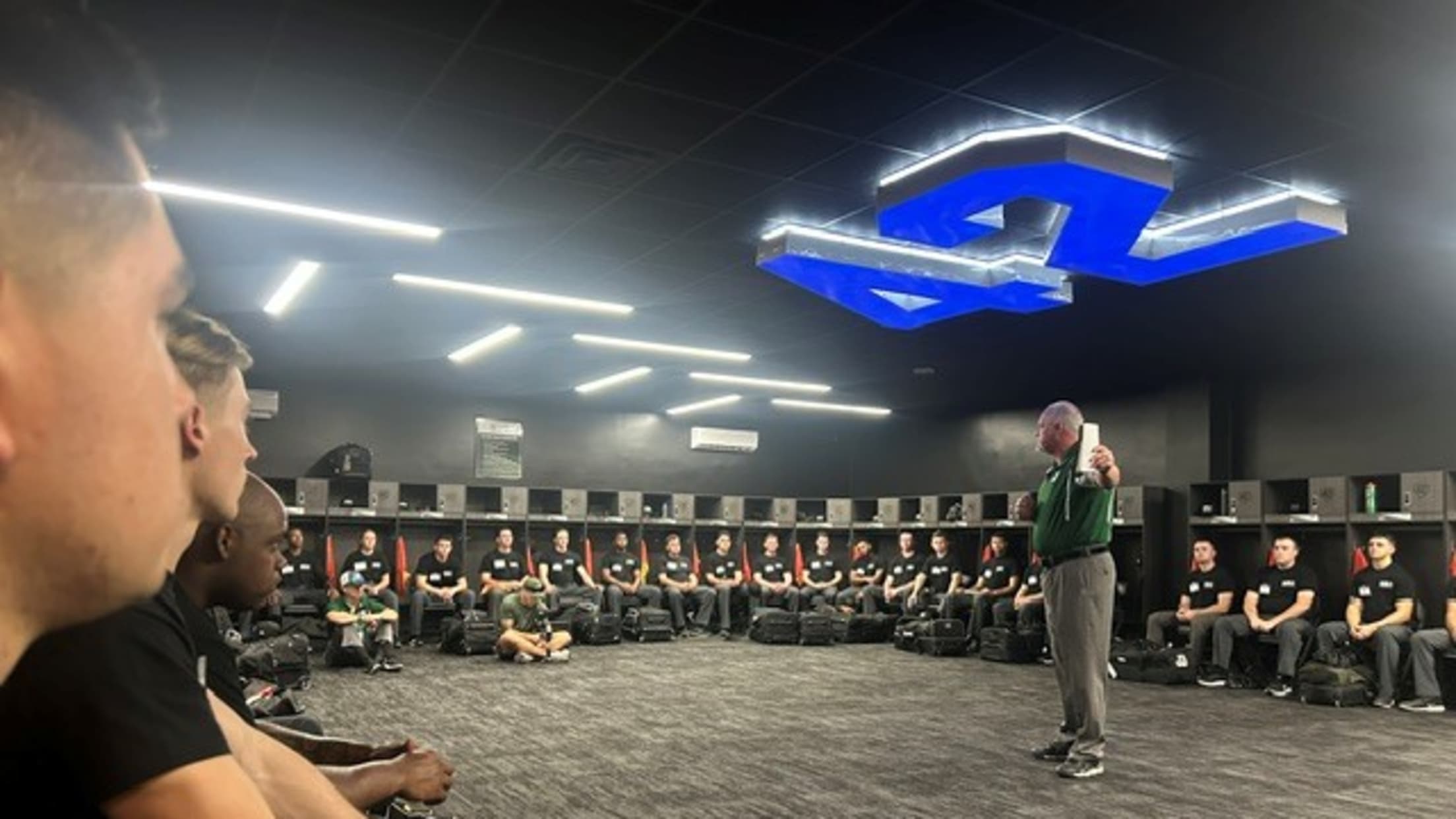 A man speaks to umpires in uniform in the locker room at the Robinson Training Complex