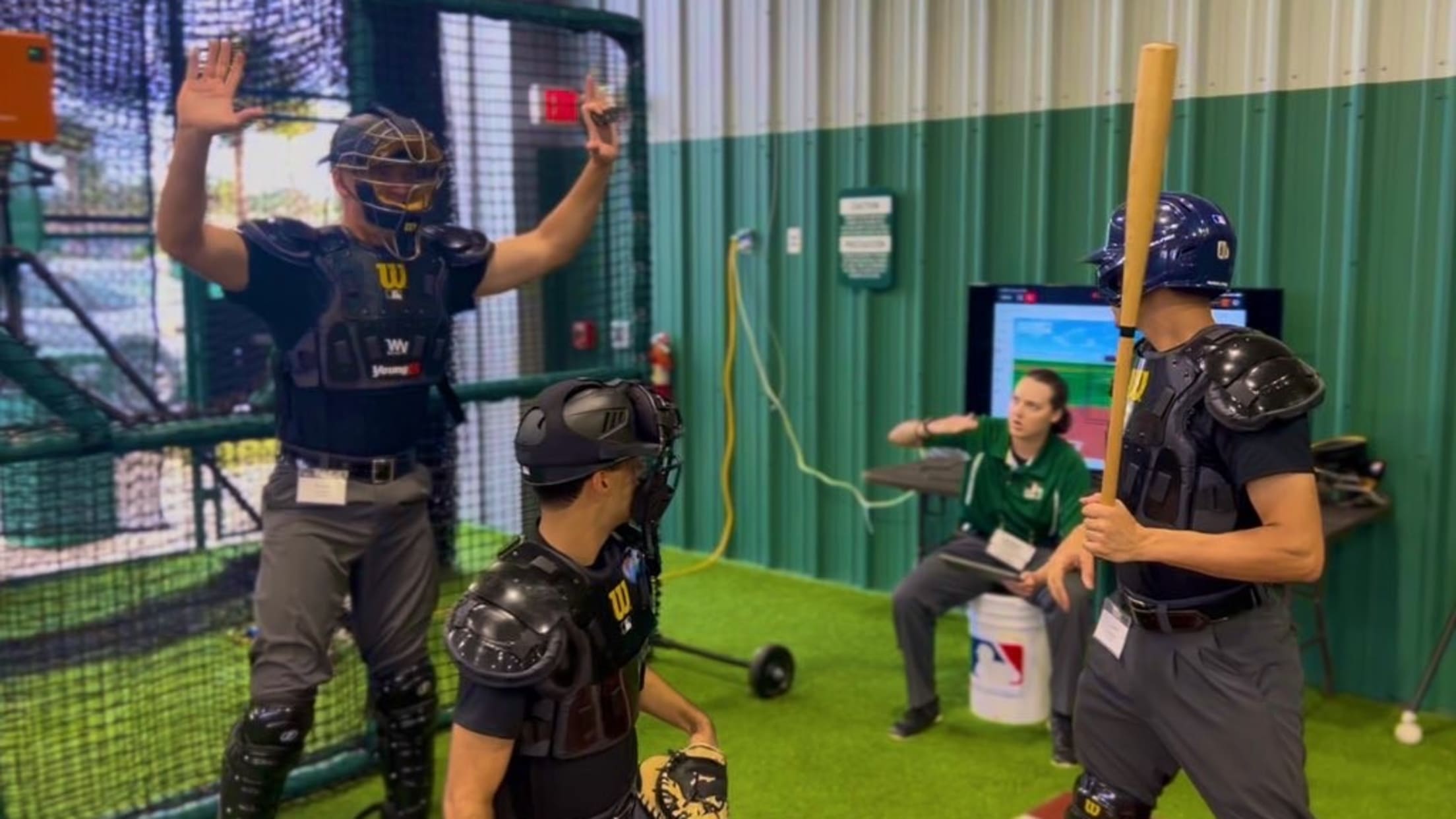An umpire prospect holds his hands up while fellow prospects in a catcher and batter position listen to a person in a green polo.