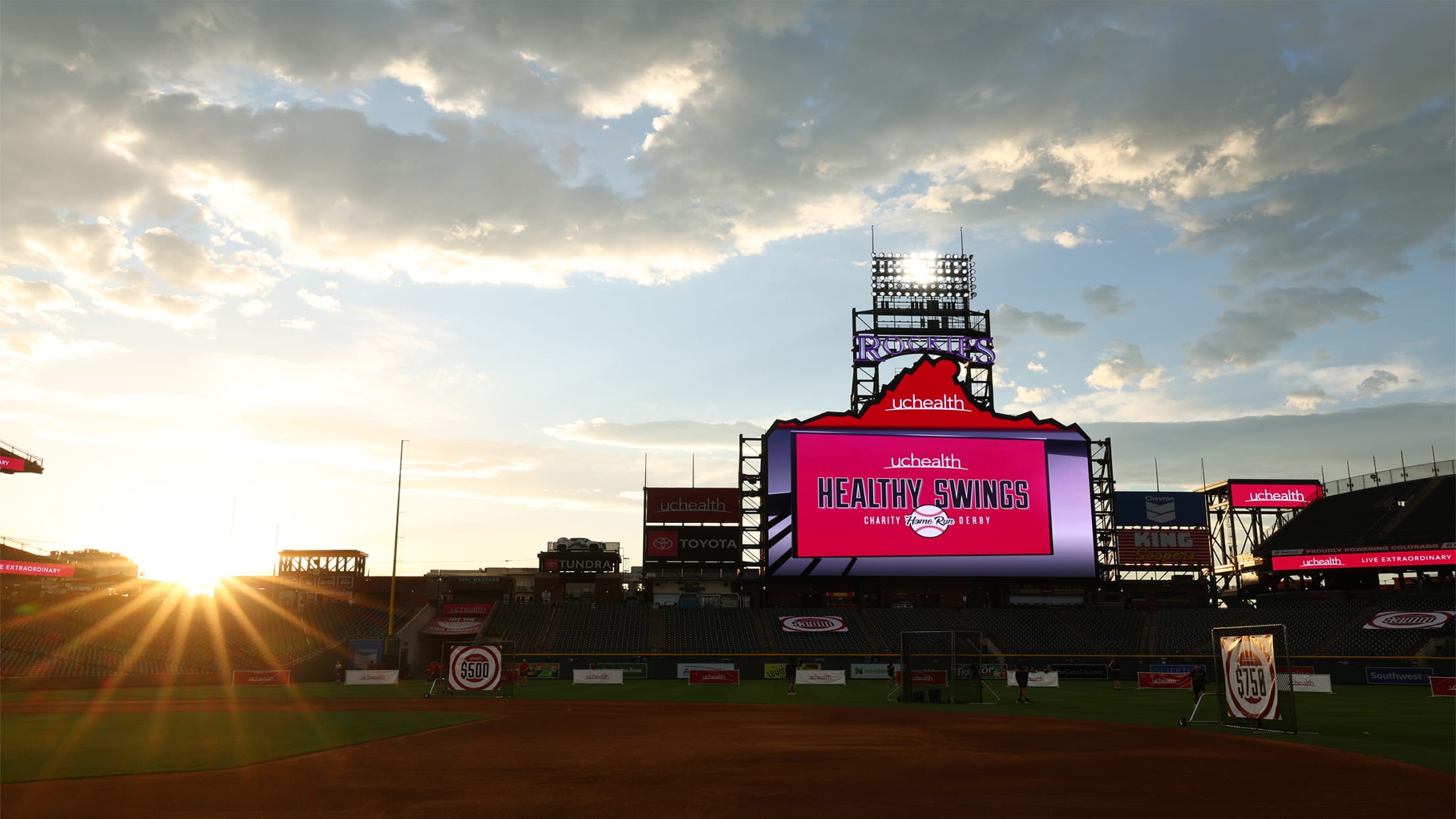 Sunset over Coors Field in Denver, Colorado during UCHealth Healthy Swings Charity Home Run Derby, with stadium seats, large digital scoreboard, and Rockies signage