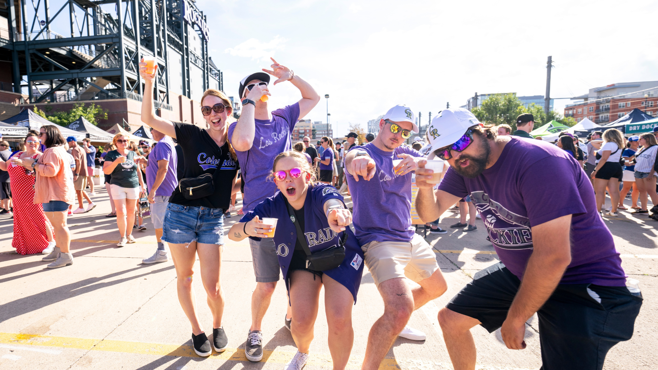 Five Rockies fans in purple shirts and jerseys smiling and pointing at the camera with people in the background drinking from plastic cups