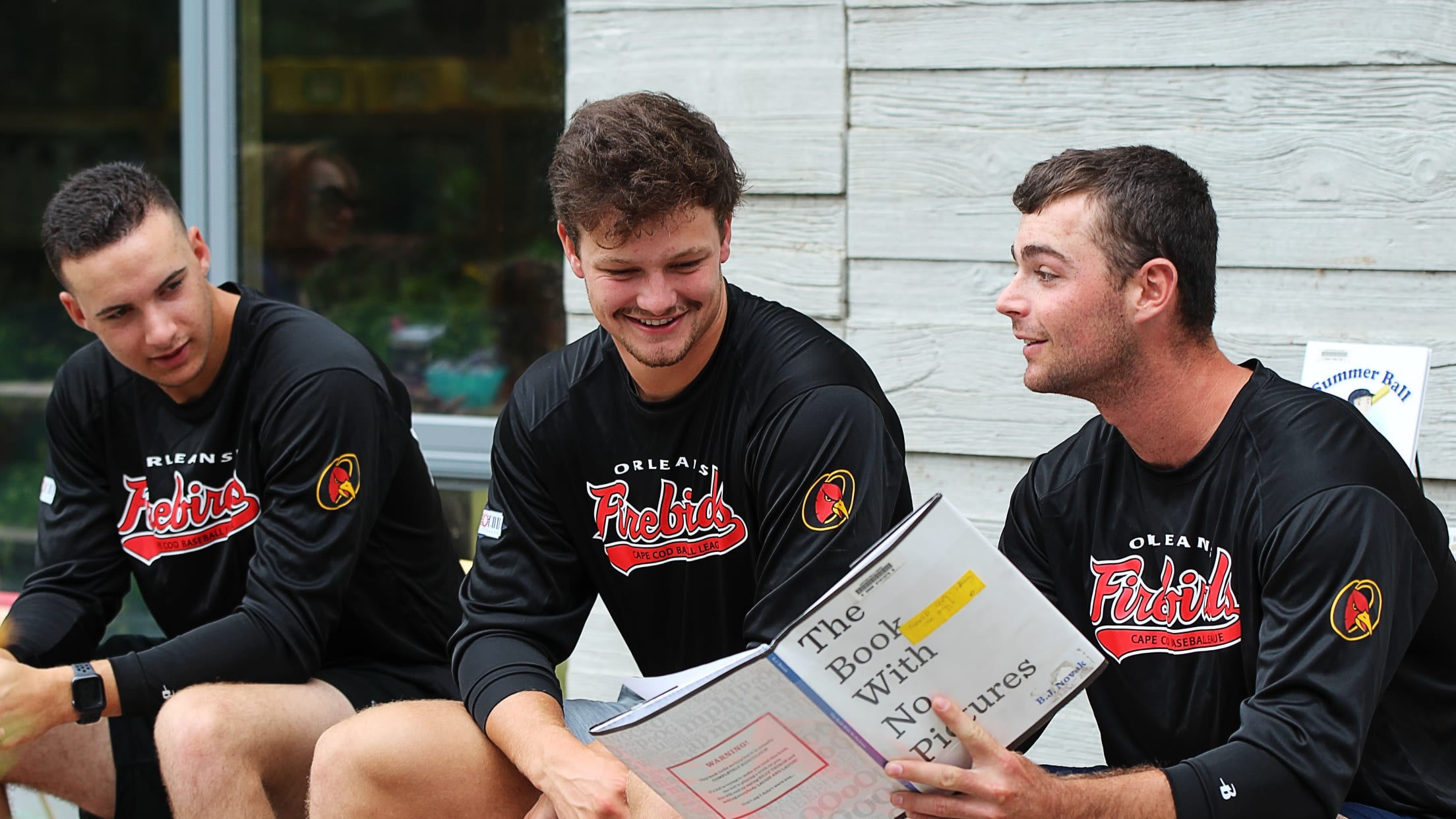 Three Firebirds players sit outside reading a book (The Book With No Pictures) to children
