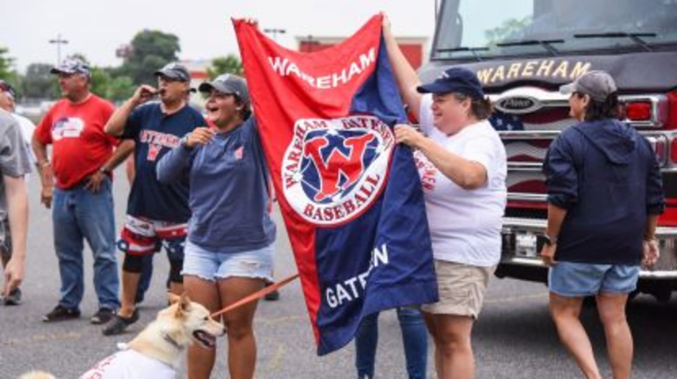 Wareham fans (including a delightful dog!) stand in the parking lot, supporting the team and waving a Wareham Gatemen flag.