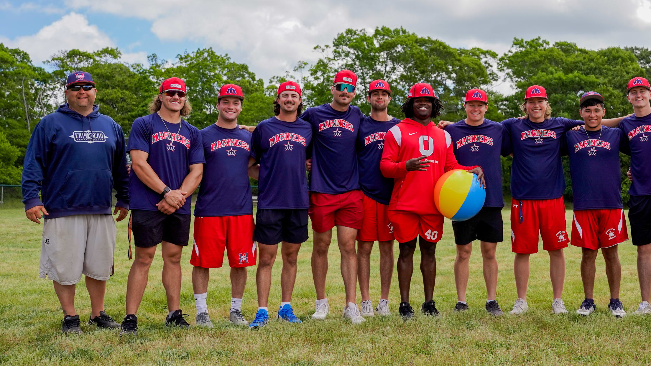 Harwich Mariners players and a coach stand in the outfield smiling for a photo during a youth clinic. Player in the middle is holding a beach ball.