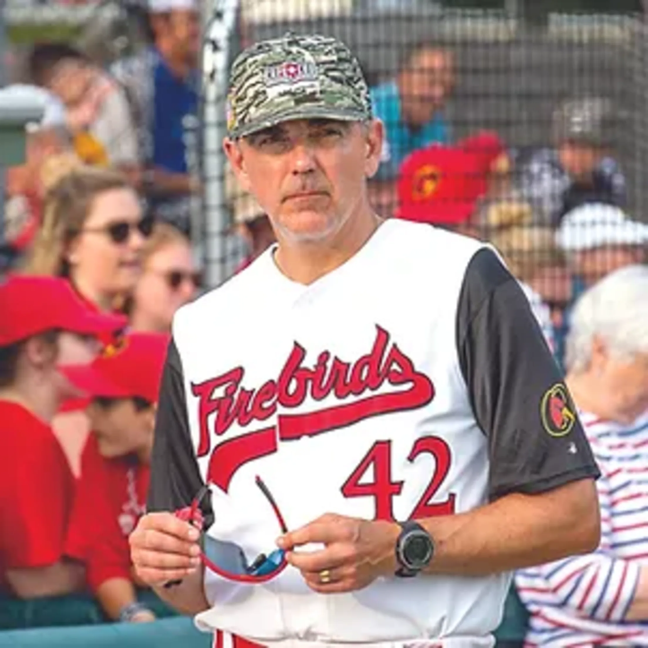 Orleans coach Jay Banfield stands while wearing his No. 42 uniform and holding his sunglasses in his hands.