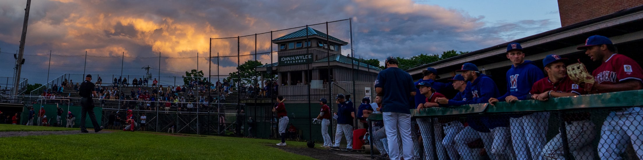 The sun begins to set above Spillane Field as the Gatemen players stand in the dugout, preparing to play.