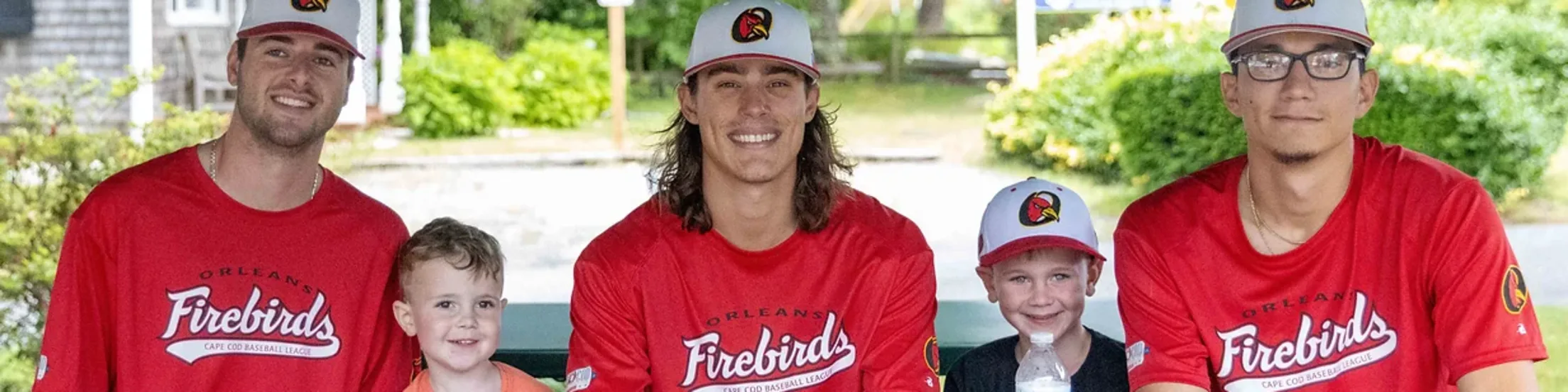 Three Orleans Firebirds players sit with two children at a table with baseball caps and Firebirds programs.