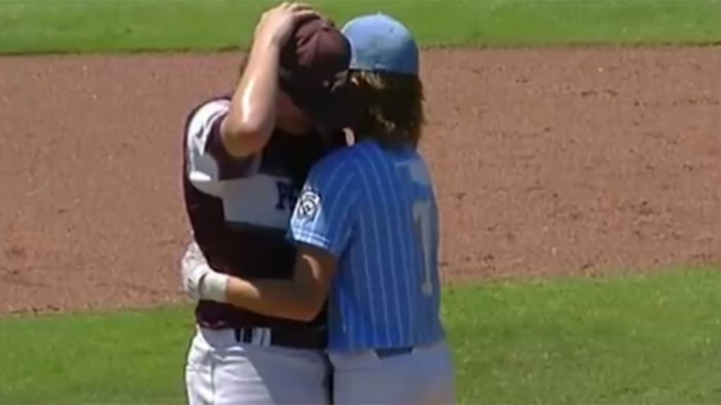 A player in a blue cap and jersey hugs a player in a maroon red cap and jersey in a Little League game