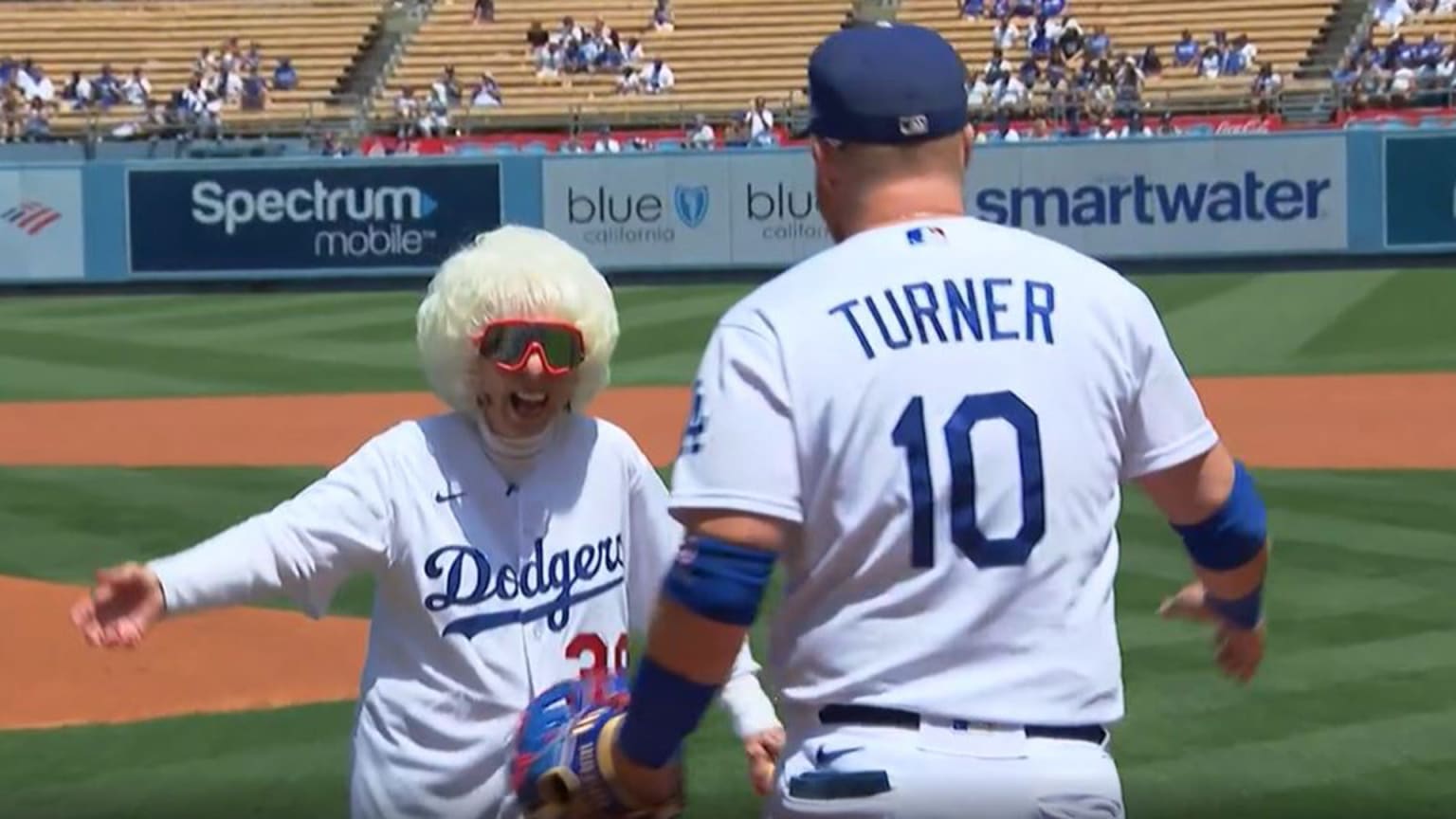 A 95-year-old woman with white hair wearing sunglasses smiles and approaches the Dodgers' Justin Turner (seen from behind) for a hug after throwing the first pitch