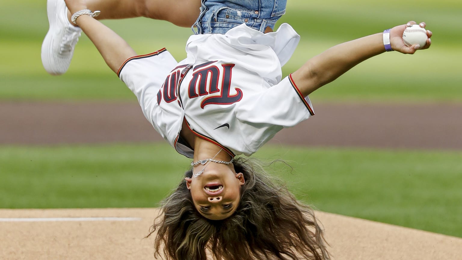 A woman in a Twins jersey upside-down, mid-flip, with a baseball in her right hand