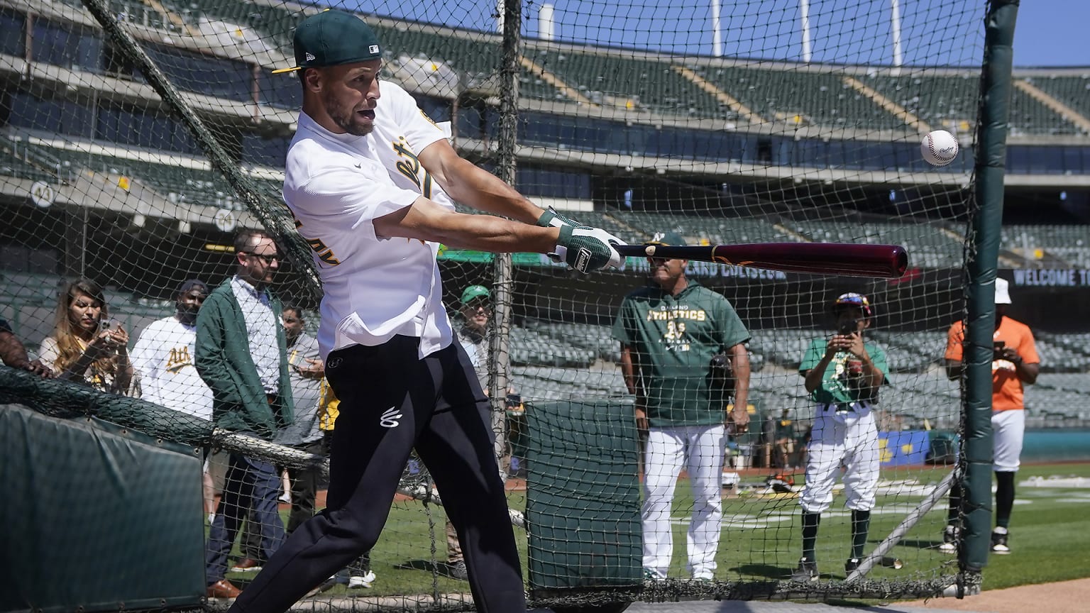 Steph Curry swings a bat in the batting cage wearing an A's jersey and cap
