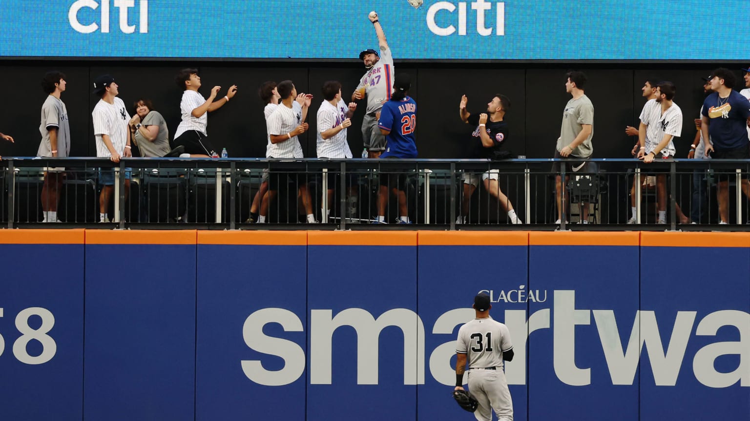 A leaping fan with a ball in his outstretched left hand stands out above other fans in a picnic area atop the outfield wall