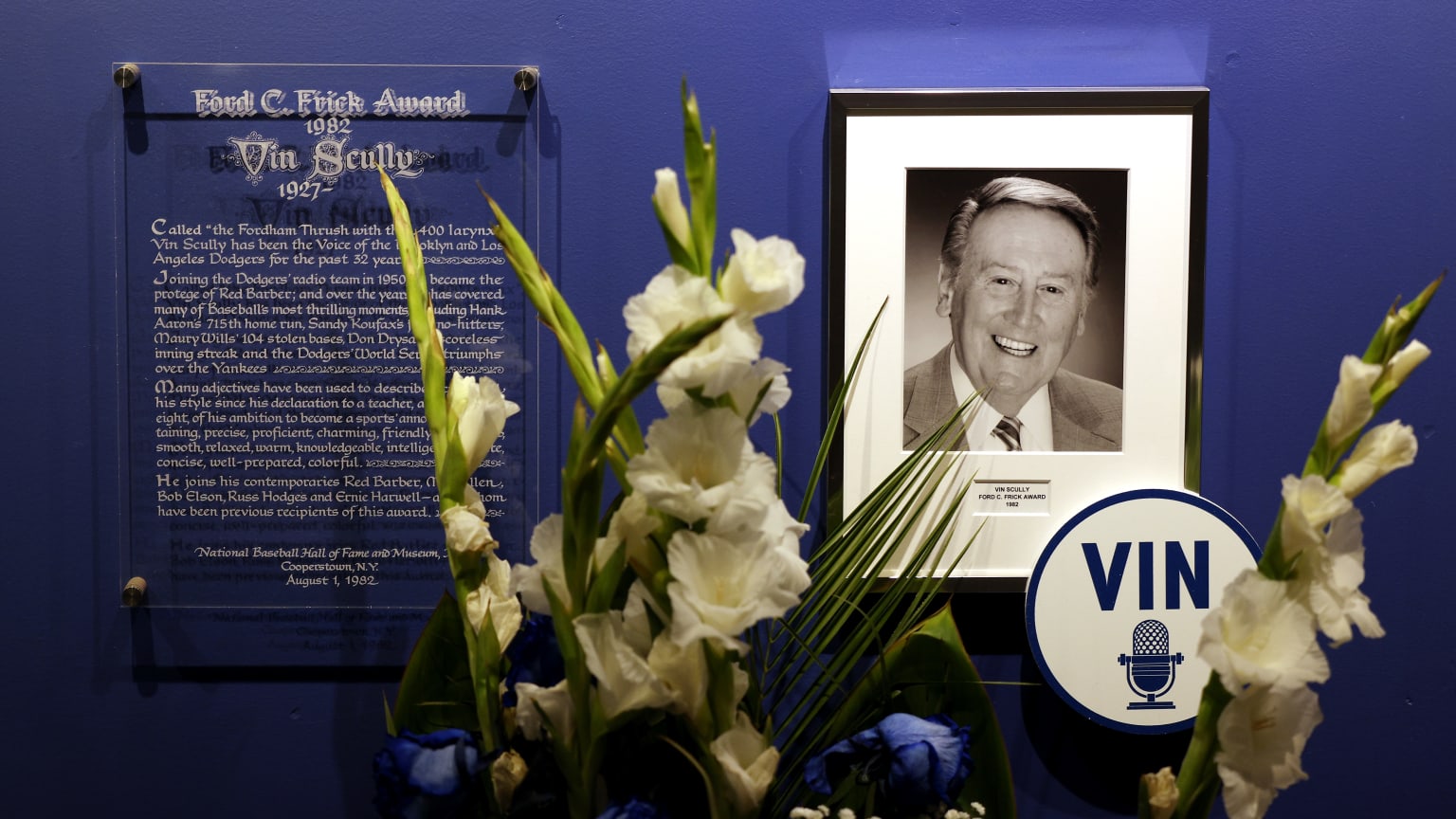 White flowers on display in front of a photograph of Vin Scully next to a plaque denoting Scully's Ford C. Frick Award recognition
