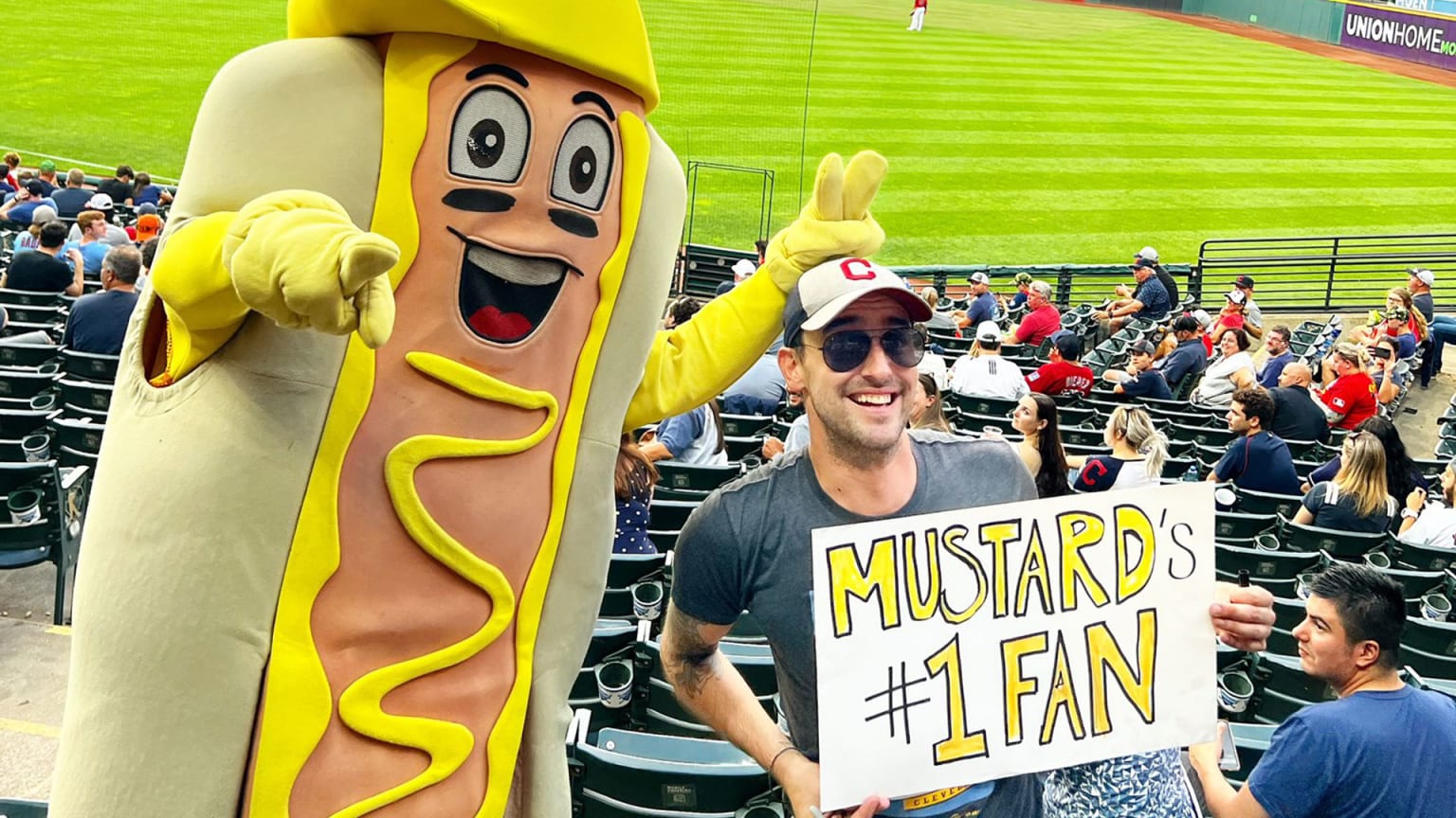 A man holds a sign that reads ''Mustard's number 1 fan''; next to him is a hot dog mascot slathered in mustard