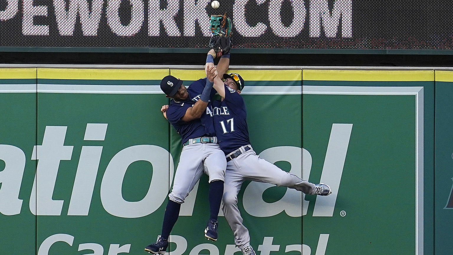 Two Mariners outfielders collide as they leap against the wall, the ball hovering just above their gloves
