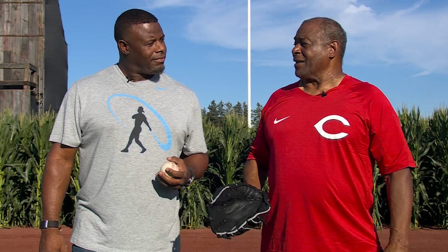 Ken Griffey Jr. in a gray T-shirt holds a baseball and looks at his dad, wearing a Reds T-shirt and holding a glove