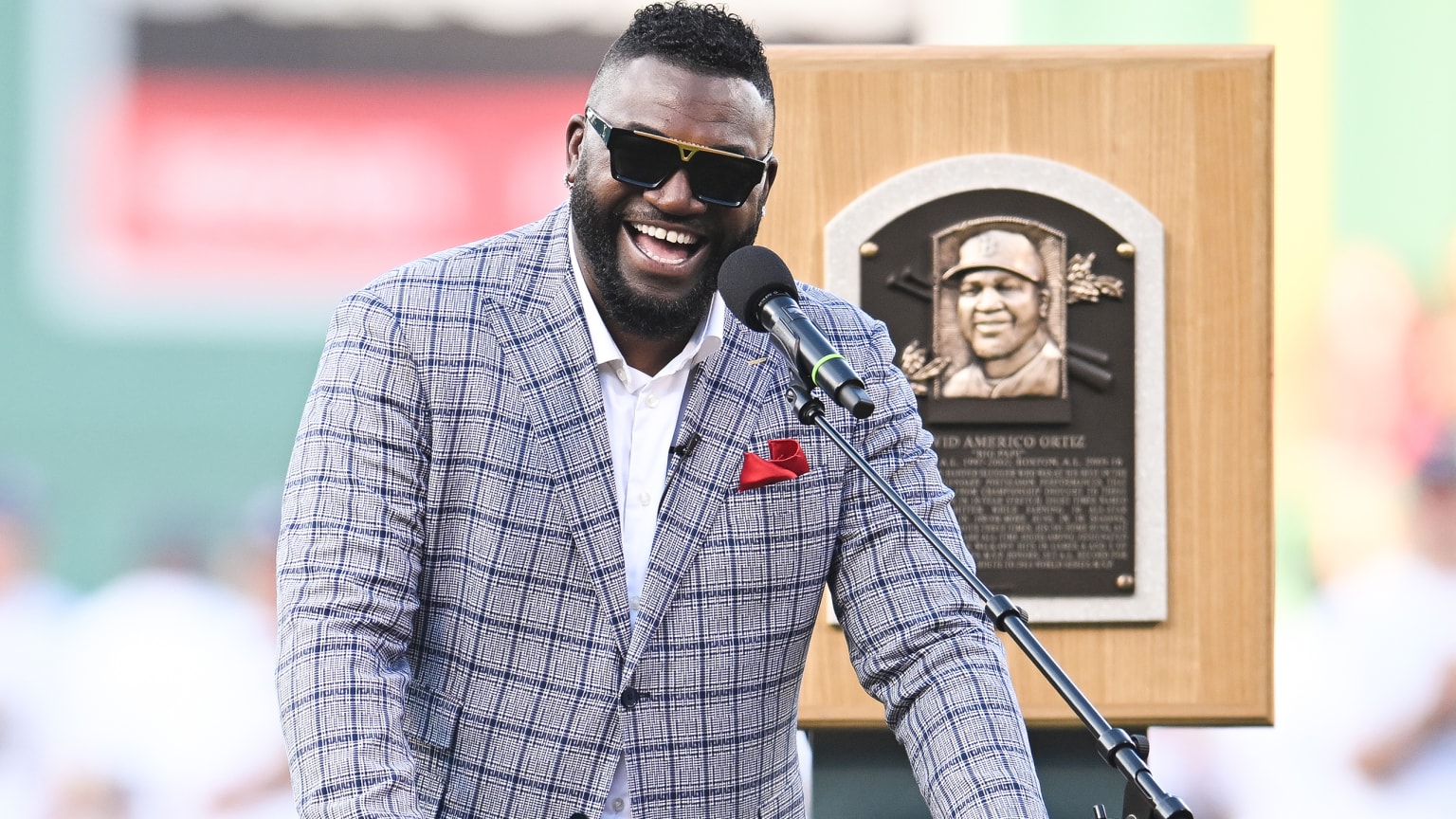 David Ortiz smiles in front of a microphone on the field, with his Hall of Fame plaque mounted behind him