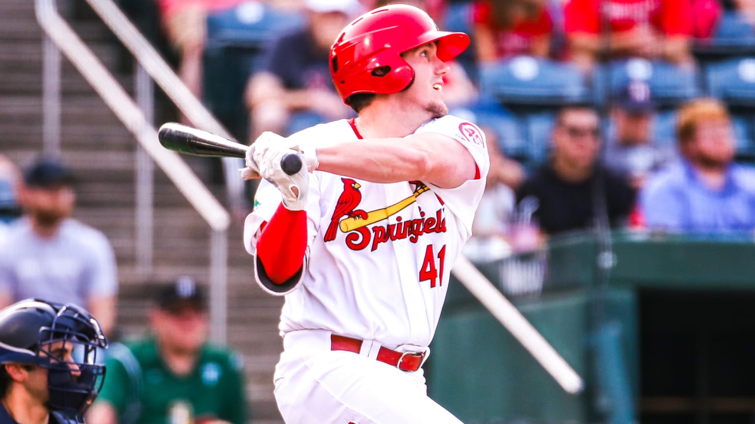 A batter in a red helmet and white uniform reading ''Springfield'' follows through on a swing