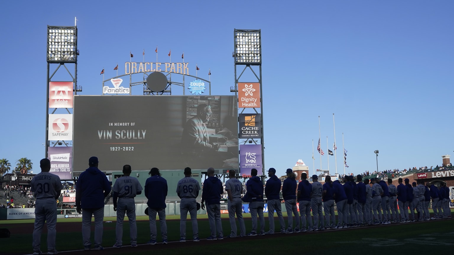 Players stand in shadow as the Oracle Park scoreboard shows an image of Vin Scully