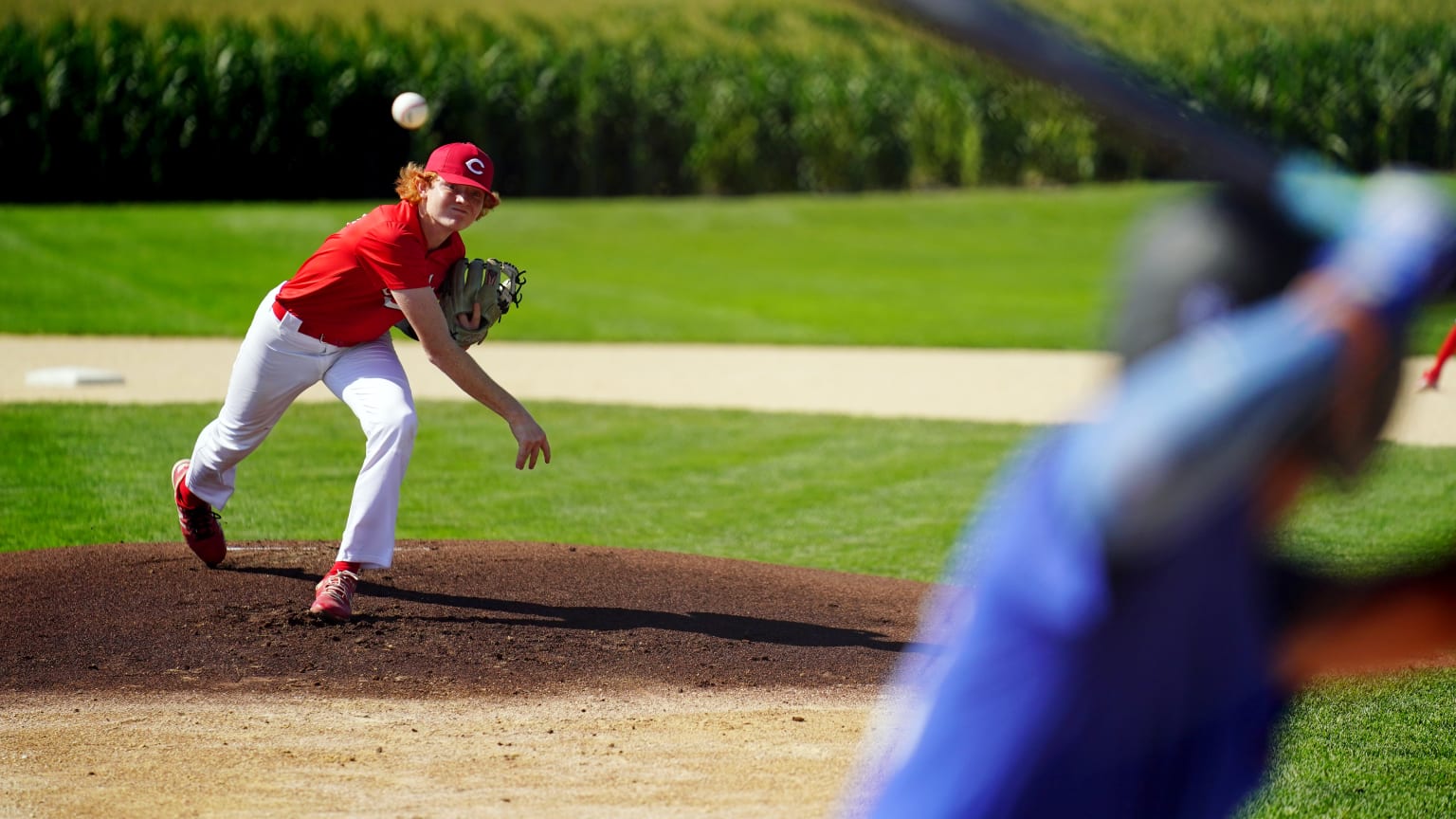 A youth pitcher in a red jersey throws from the mound at the Field of Dreams to a batter in blue in the foreground