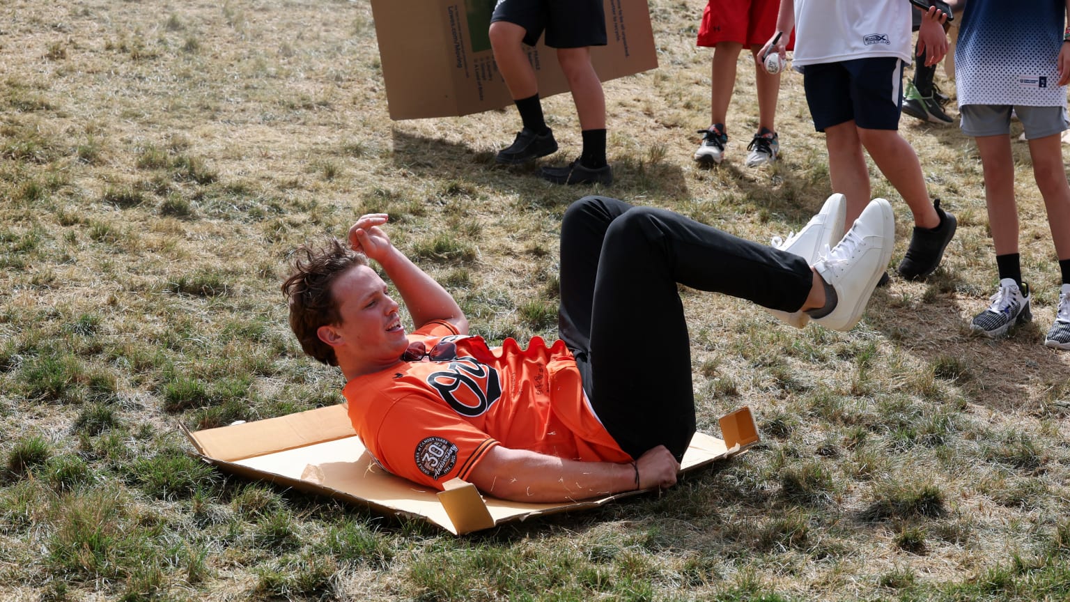 A player in an orange Orioles jersey lies on his back on a piece of cardboard, feet in the air as he slides down a grassy hill