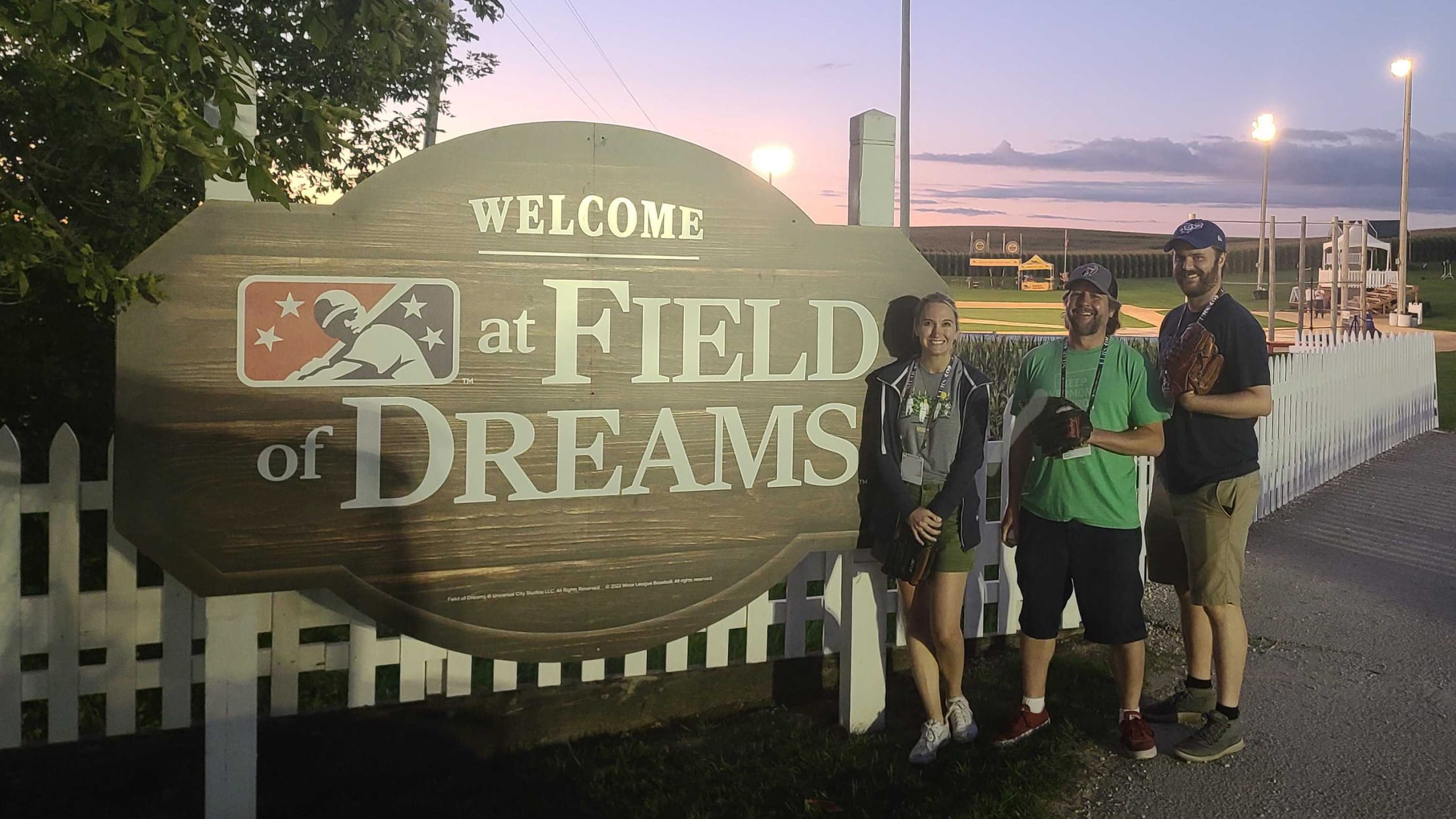 A woman and two men stand next to a large wooden sign reading, ''Welcome: Minor League Baseball at Field of Dreams''