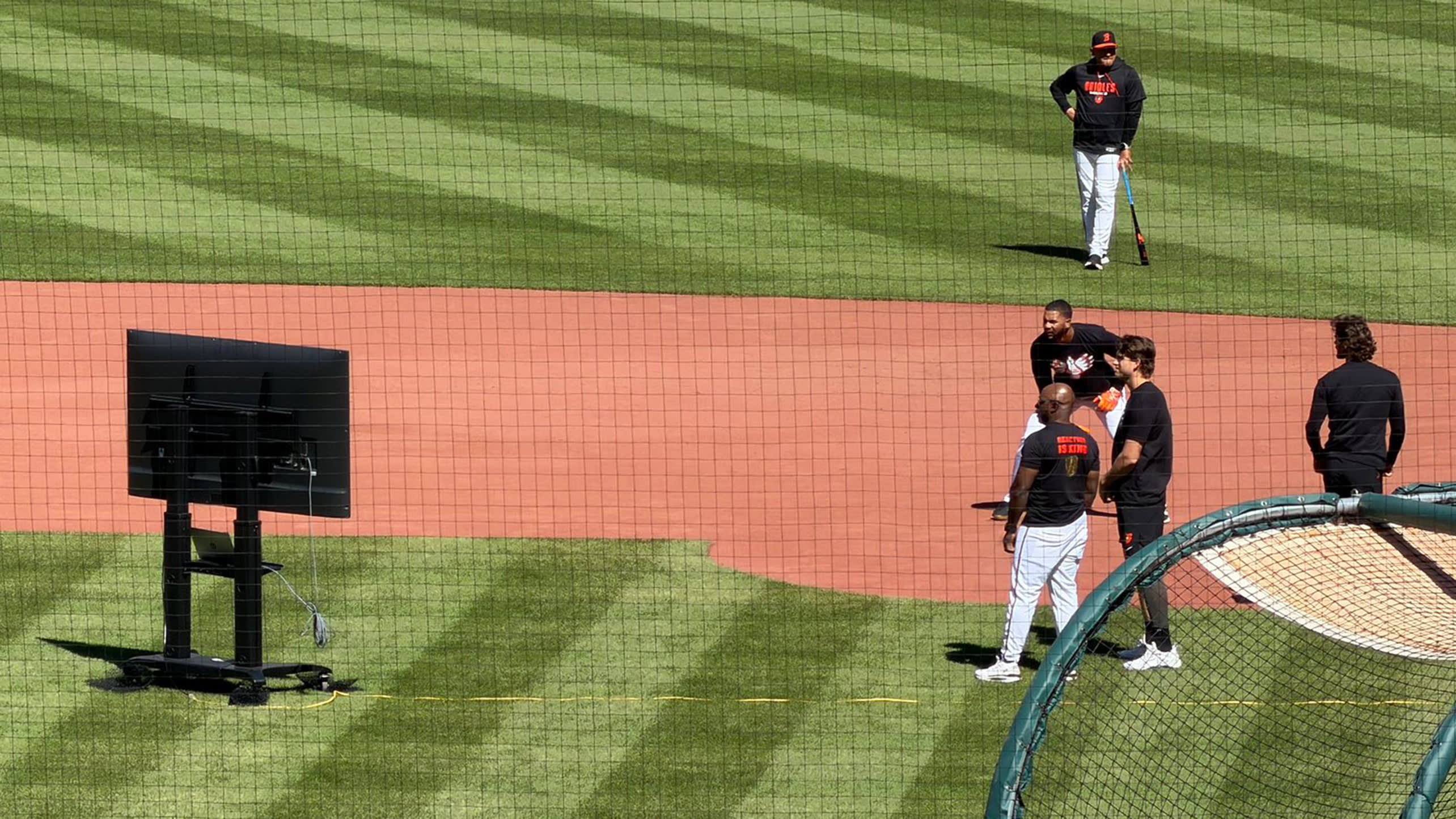 Orioles players and coaches look at an 80-inch TV on the infield grass