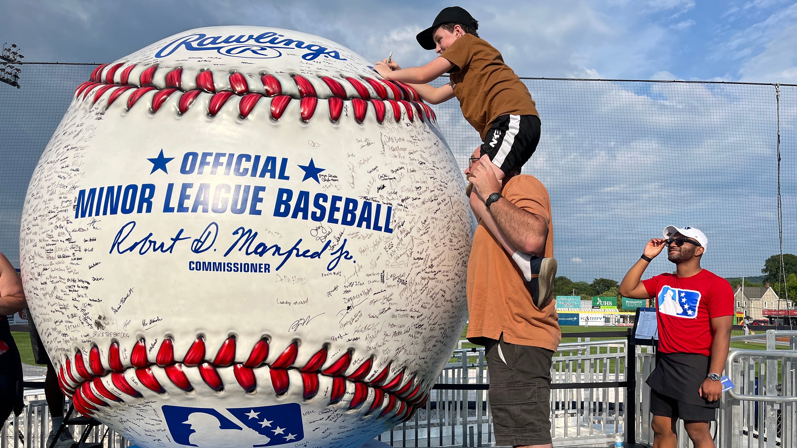 A child on someone's shoulders signs a giant baseball