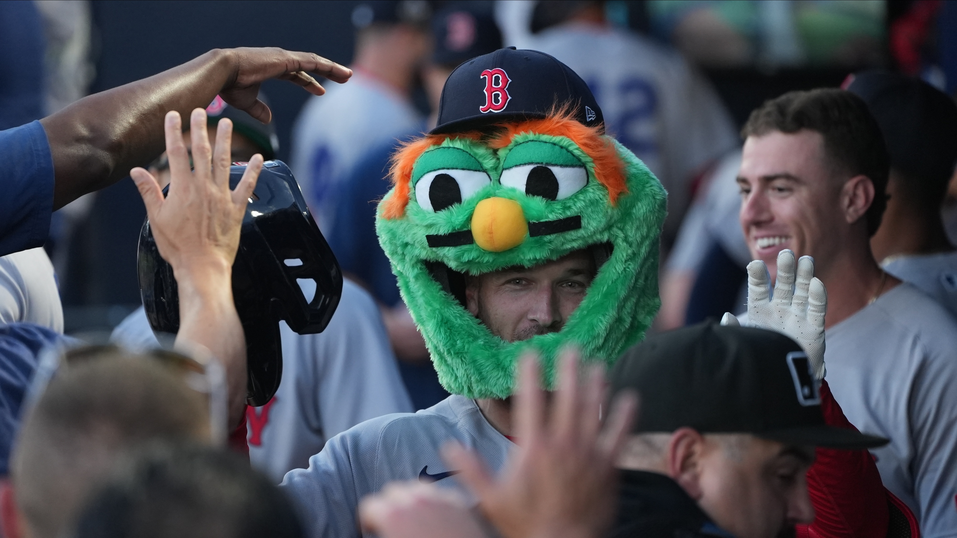 Alex Bregman, wearing the head of a green, fuzzy mascot with a Red Sox cap atop it, receives high-fives in the dugout