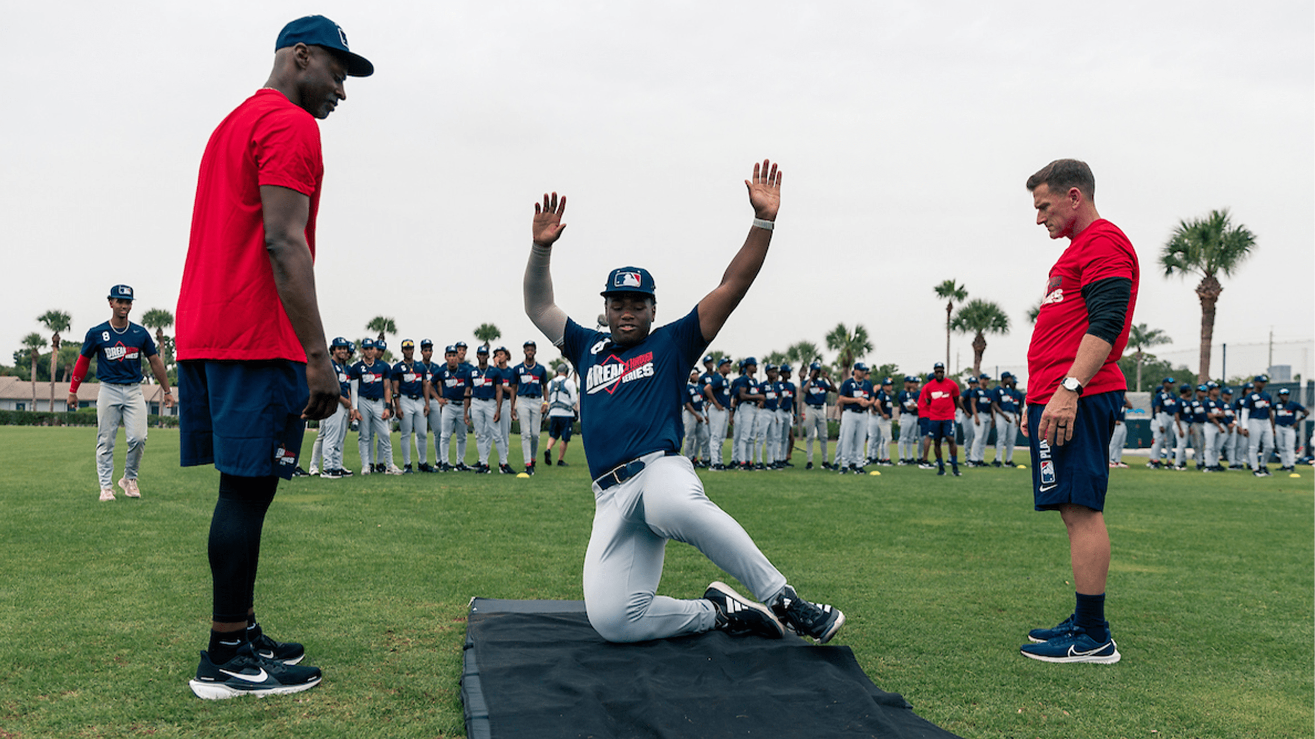 A high school player slides onto a mat in a drill as two coaches look on and his peers line up behind him