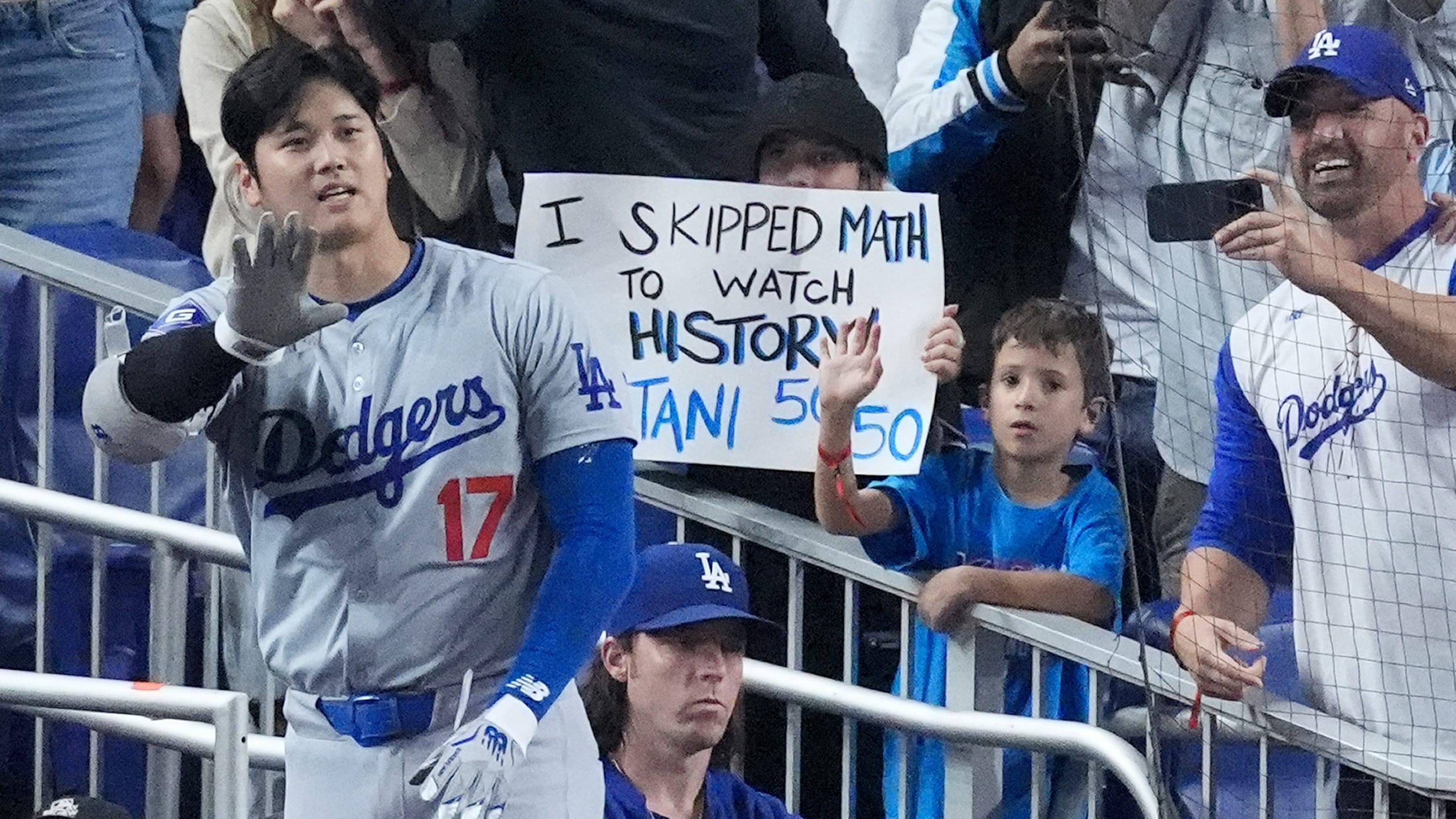 Shohei Ohtani waves from the top step of the dugout in front of a fan holding a sign