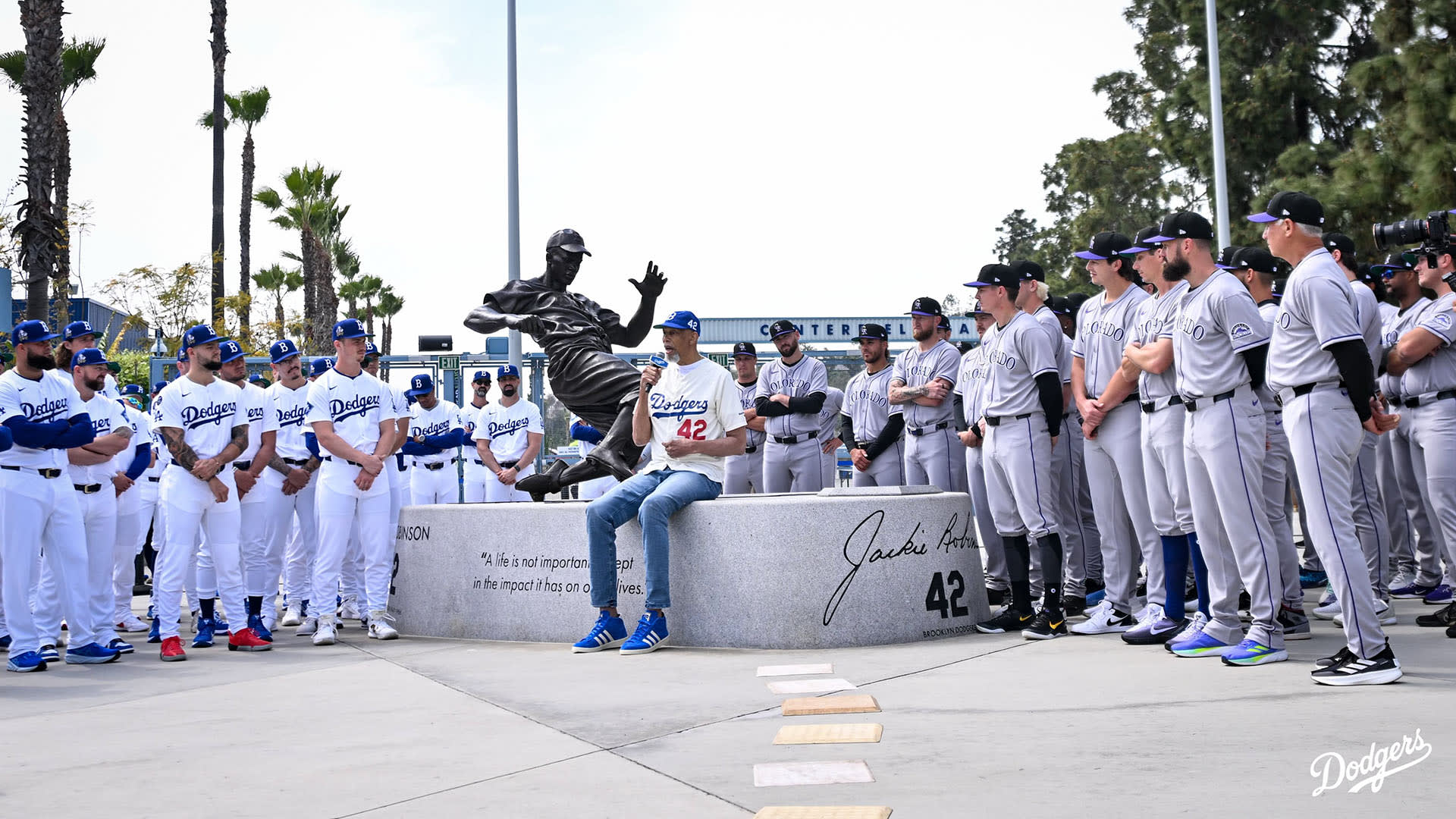NBA Hall of Famer Kareem Abdul-Jabbar wears a Jackie Robinson jersey while sitting in front of his statue at Dodger Stadium. Uniformed Rockies players stand to the right and Dodgers players in uniform stand on the left