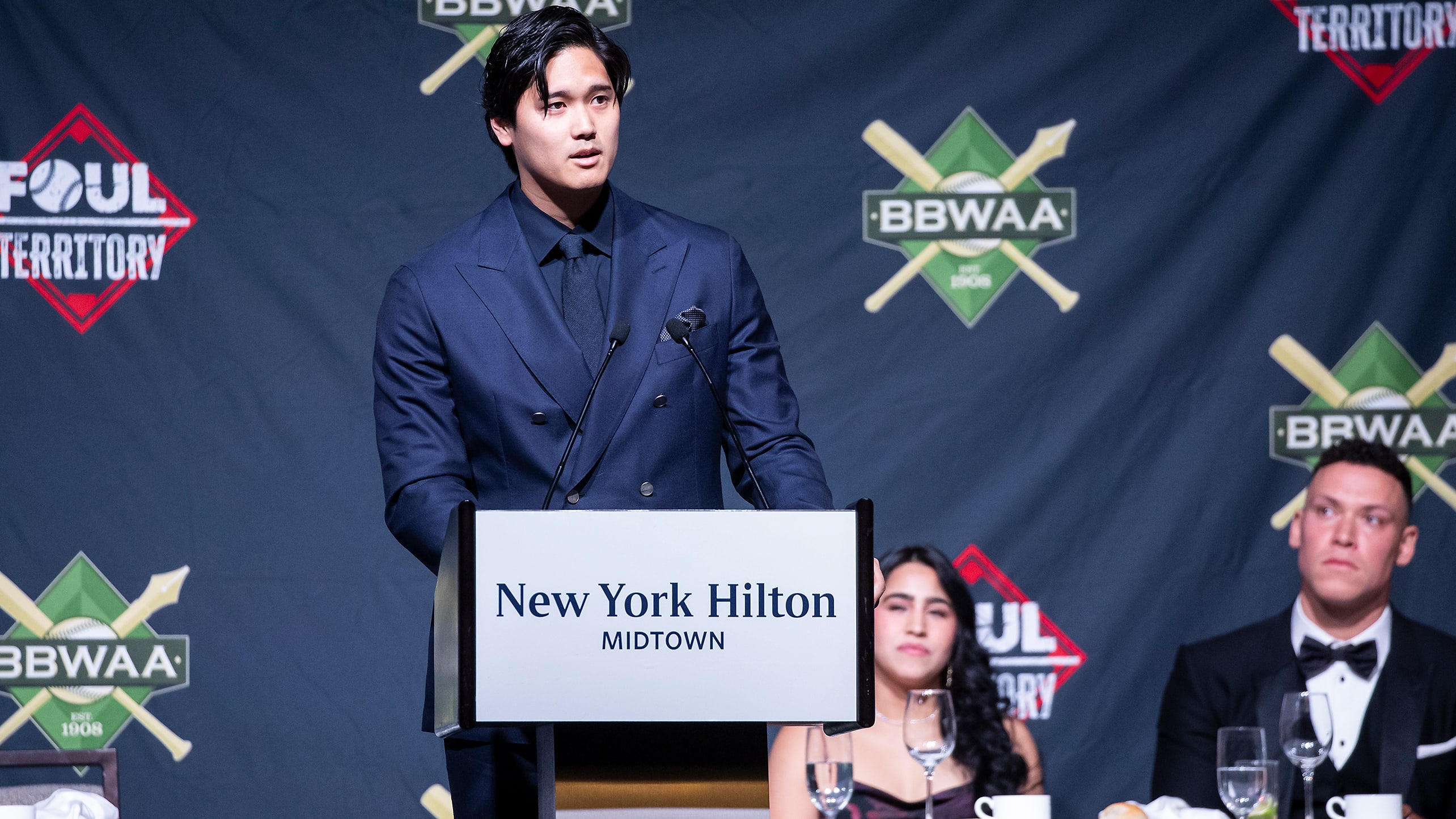 Shohei Ohtani speaks at a podium as Aaron Judge looks on