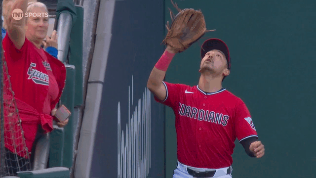 An animated gif of Steven Kwan making a catch and a fan in the stands waving toward the camera