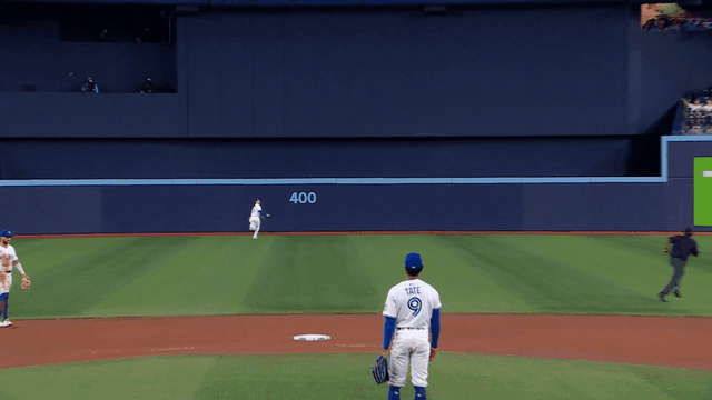 An animated gif of Daulton Varsho falling down on the warning track and making a behind-the-back catch on one knee